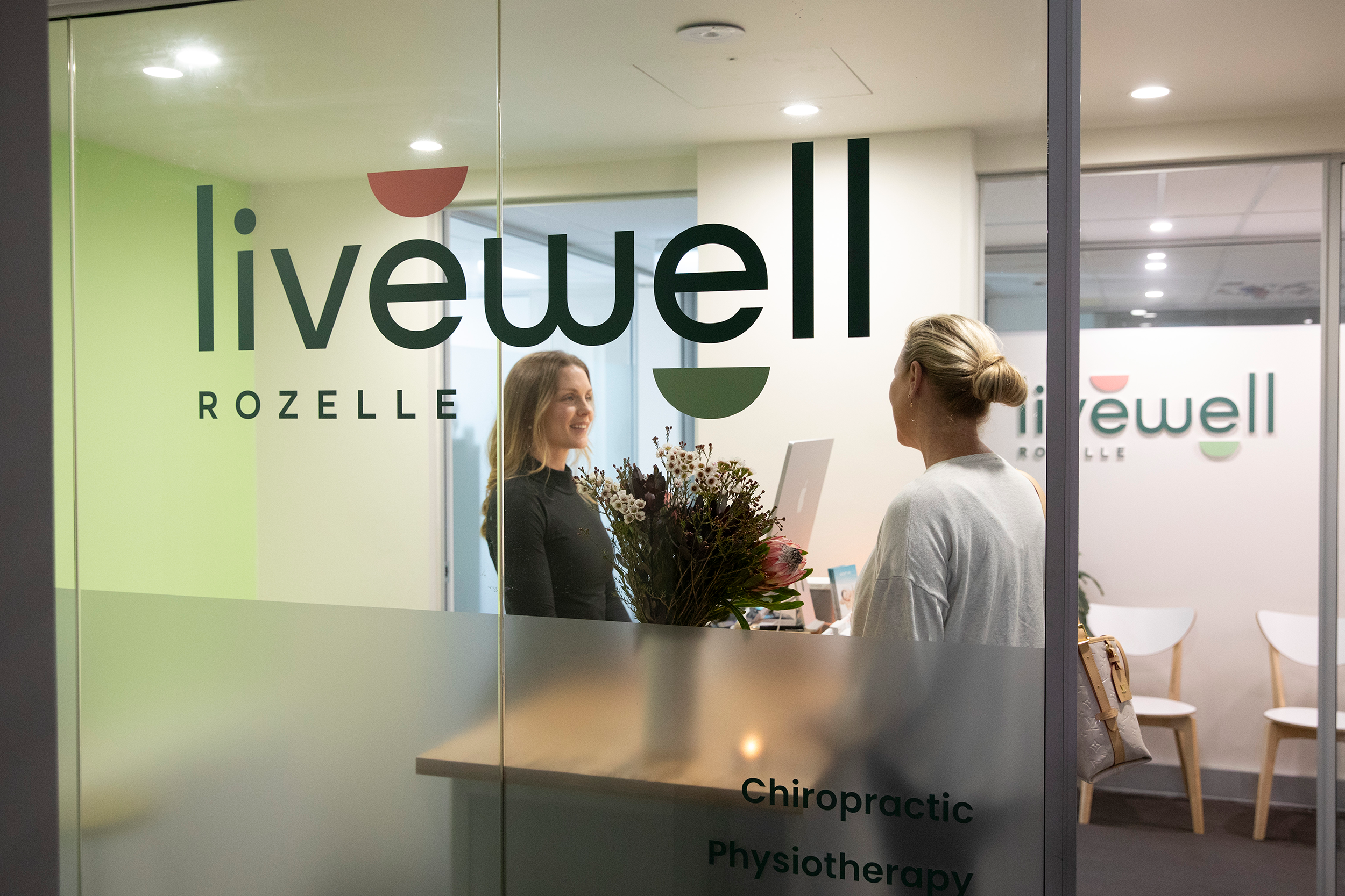 Interior of a chiropractic and physiotherapy clinic with the receptionist and client exchanging greeting, viewed through a glass wall, logo on the glass and reception chairs in background