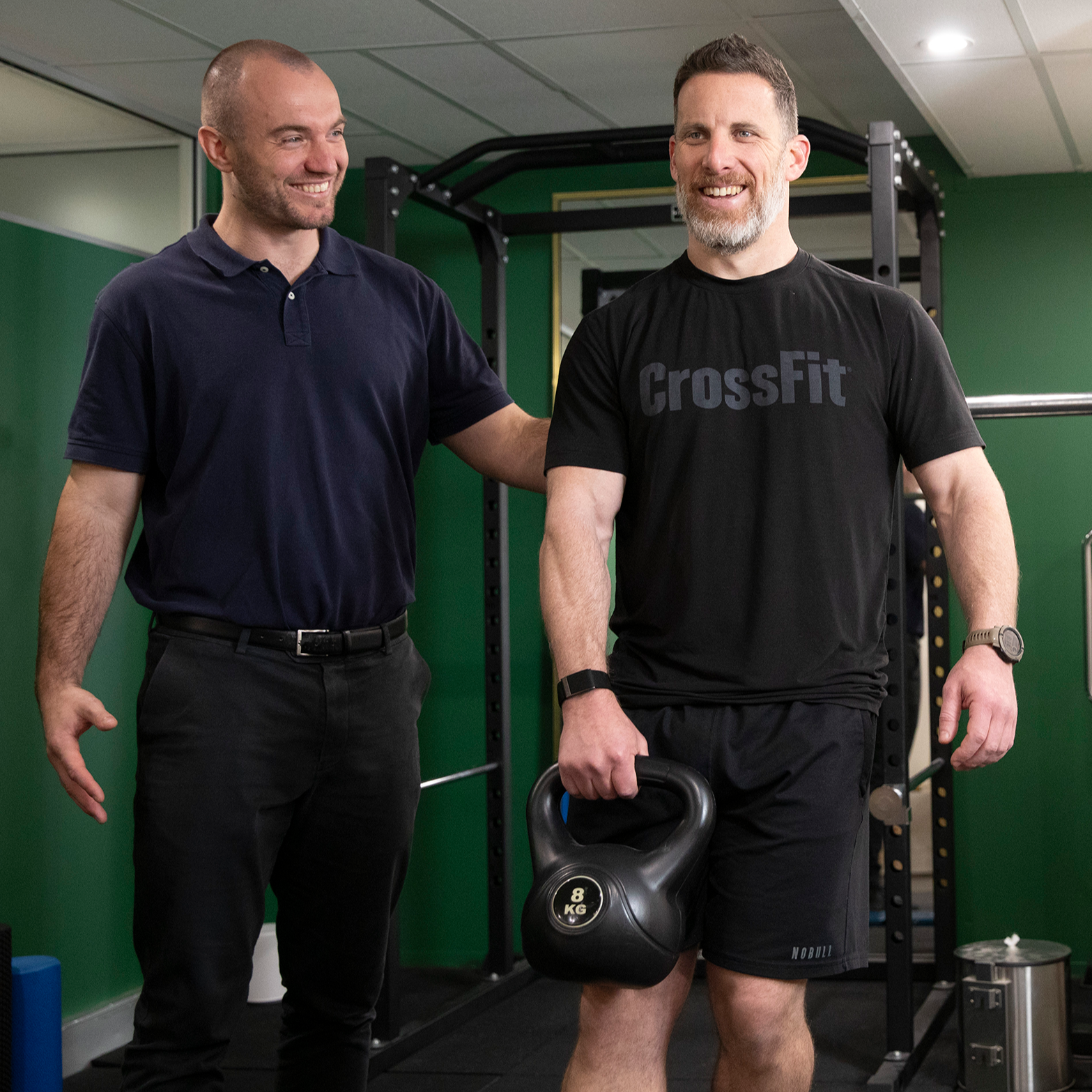 A male client in black gym gear holding a kettle bell with Chiropractic practitioner standing beside him to coach the exercise movement