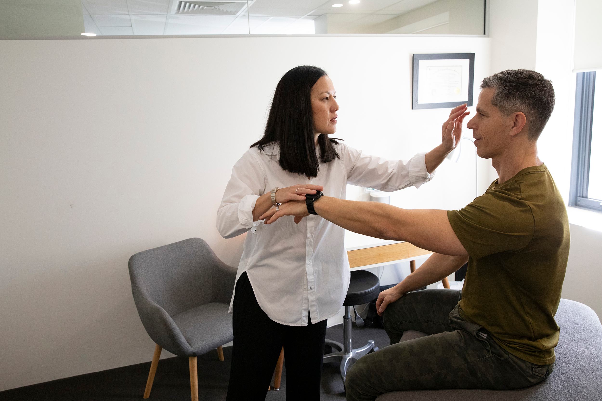 Kinesiologist standing in front of a client sitting on a treatment table. His arm is raised out in front while she pushes down to test its strength