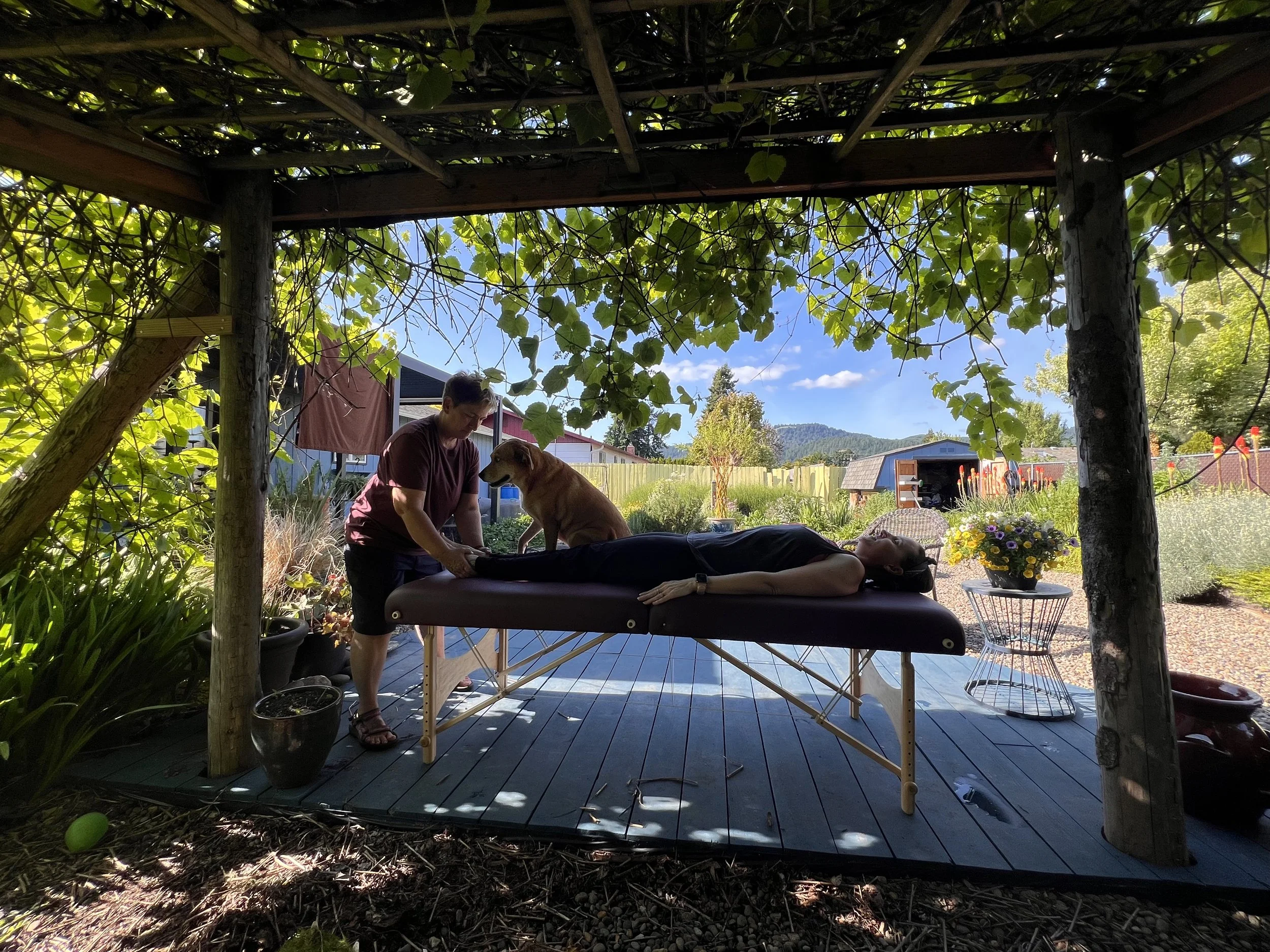 Mary Zephyr, Certified Reiki Master Teacher at Healthy Manifestations in Springfield, Oregon, offering Reiki to a client outdoors, with Zoe the dog sitting peacefully beside them. A serene moment of healing in the Eugene–Springfield community garden