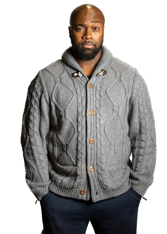 A man with a beard and mustache wearing a gray cable knit cardigan with brown buttons and a hood, standing against a black background.