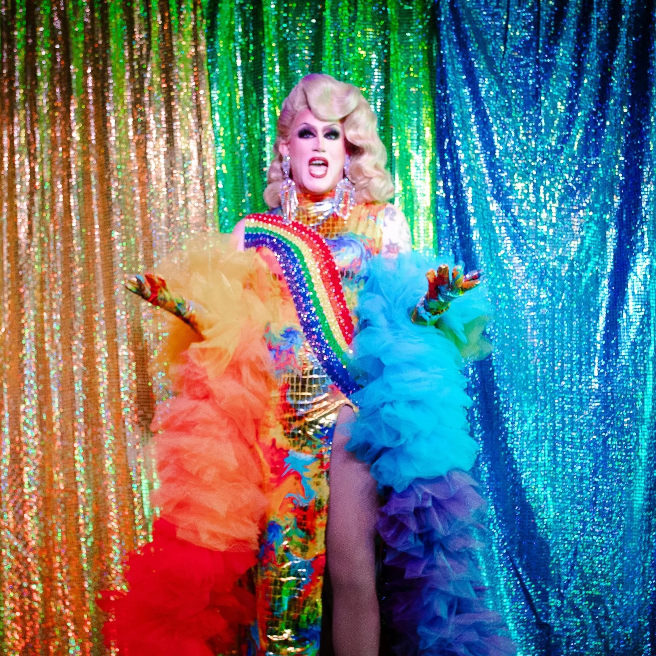 A drag performer stands against a colorful, glittery curtain, wearing a vibrant rainbow-themed outfit with rainbow jewelry and large earrings, with dramatic makeup and blonde, styled hair.