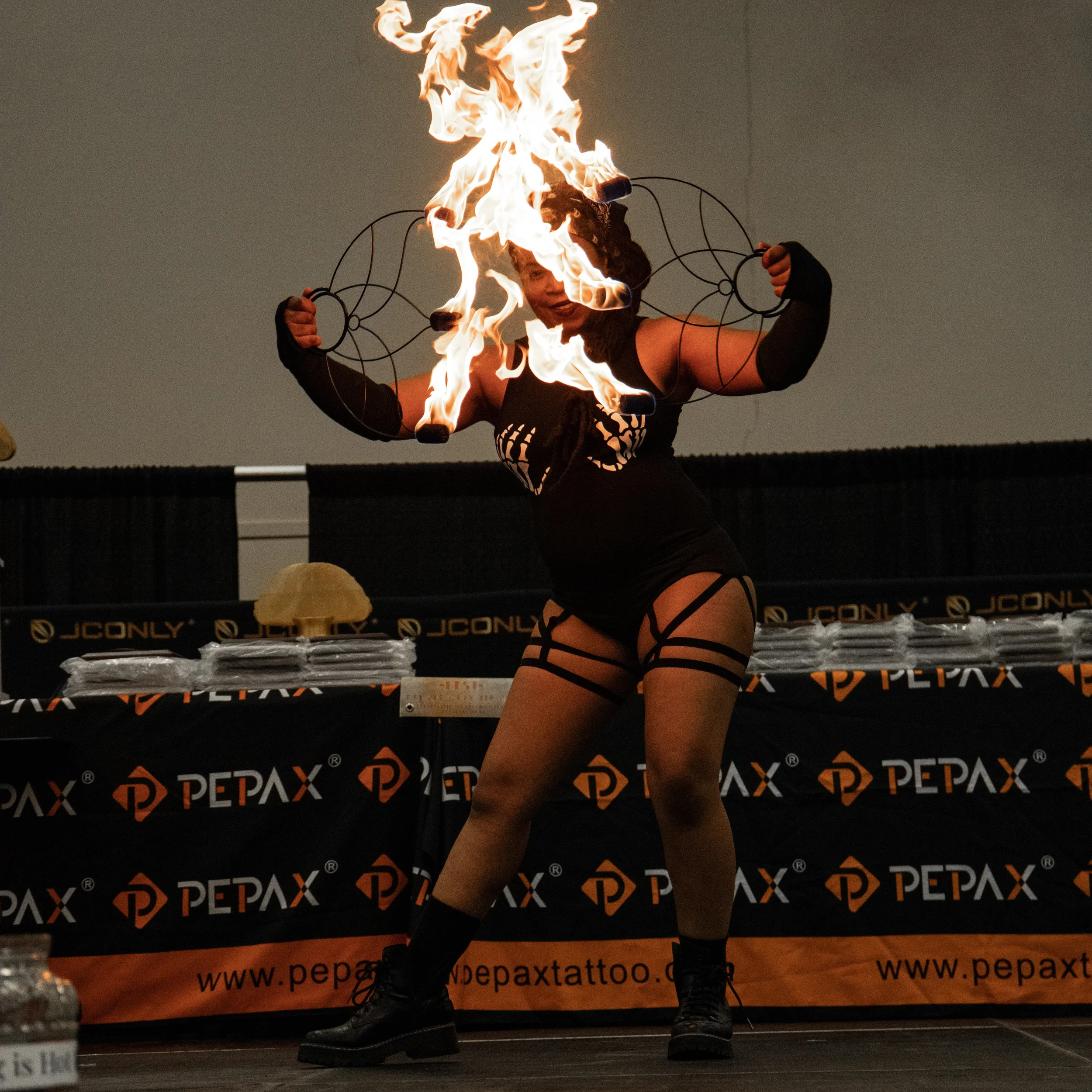 A woman performing a fire fire dancing act with a flaming torch in each hand, wearing black clothing with cutouts and arm sleeves, standing on a stage decorated with black and orange banners featuring the company PePAX.