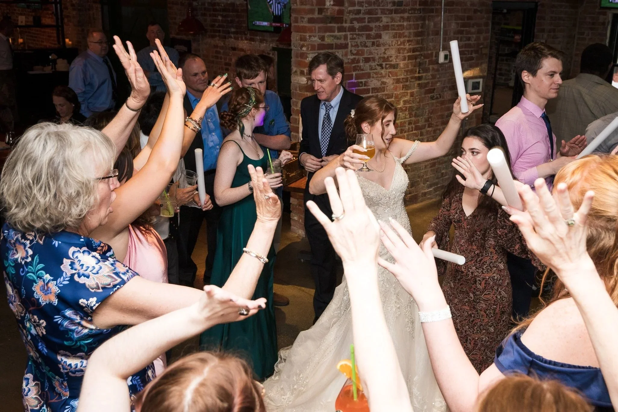 A lively wedding reception with guests dancing and celebrating, including a bride in a white gown holding a drink and surrounded by family and friends in a rustic indoor venue.