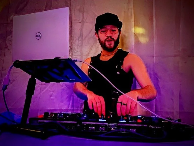 A person DJing with a laptop and DJ equipment, wearing a black tank top, glasses, a black hat, and a choker, standing in front of a purple-lit backdrop.