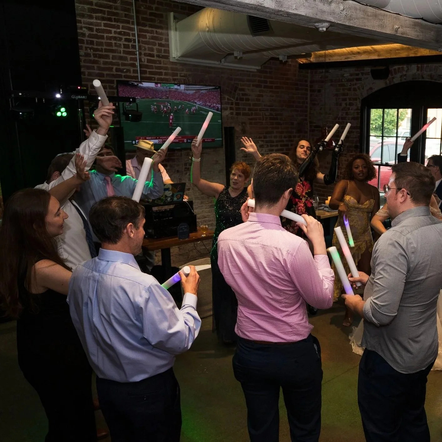 People dancing and celebrating at a party, holding LED glow sticks in a dimly lit room with a brick wall and a large window, watching a football game on a TV.