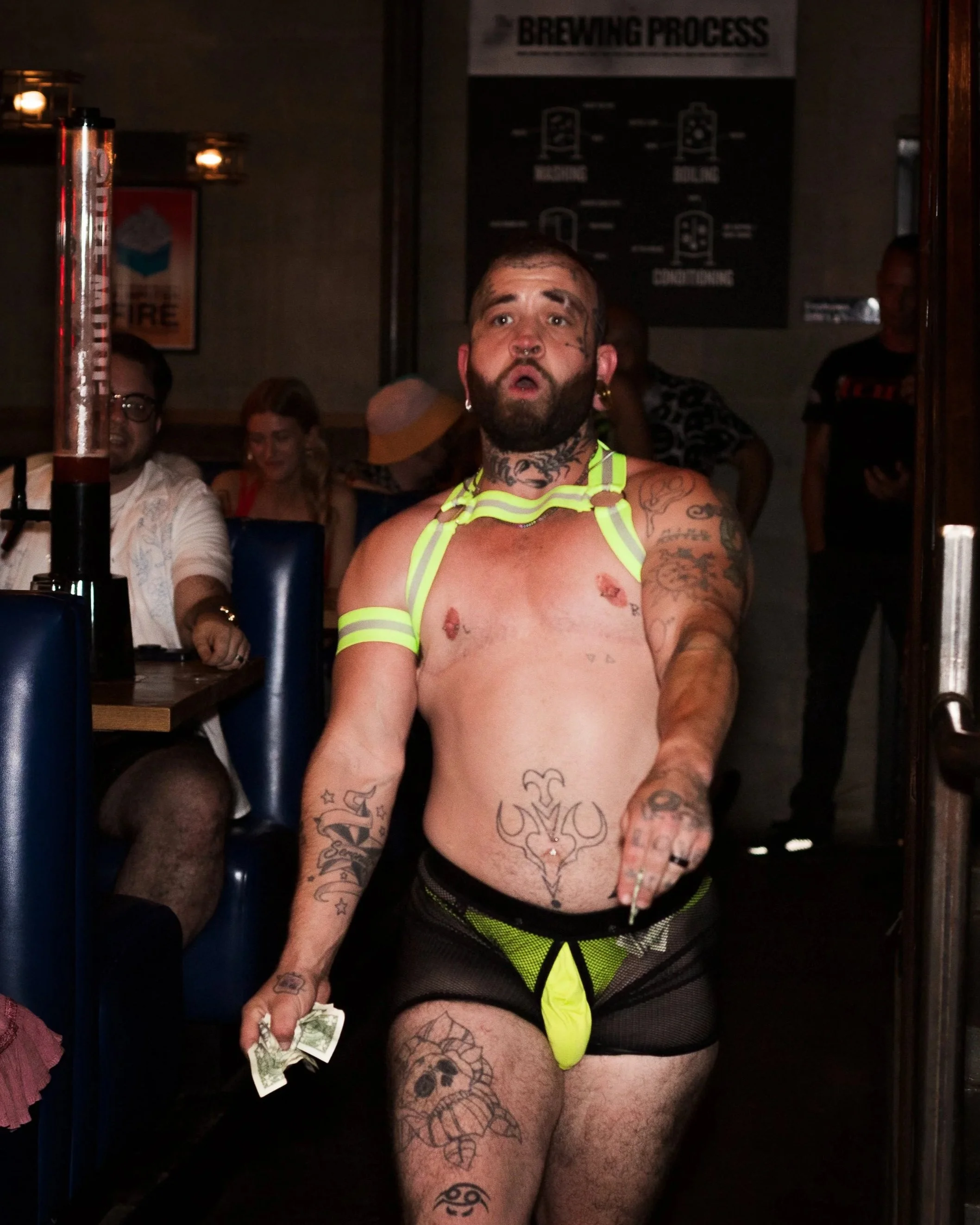 A man with tattoos and piercings wearing revealing neon green and black clothing, holding cash, standing inside a bar or club, with other people seated in the background.