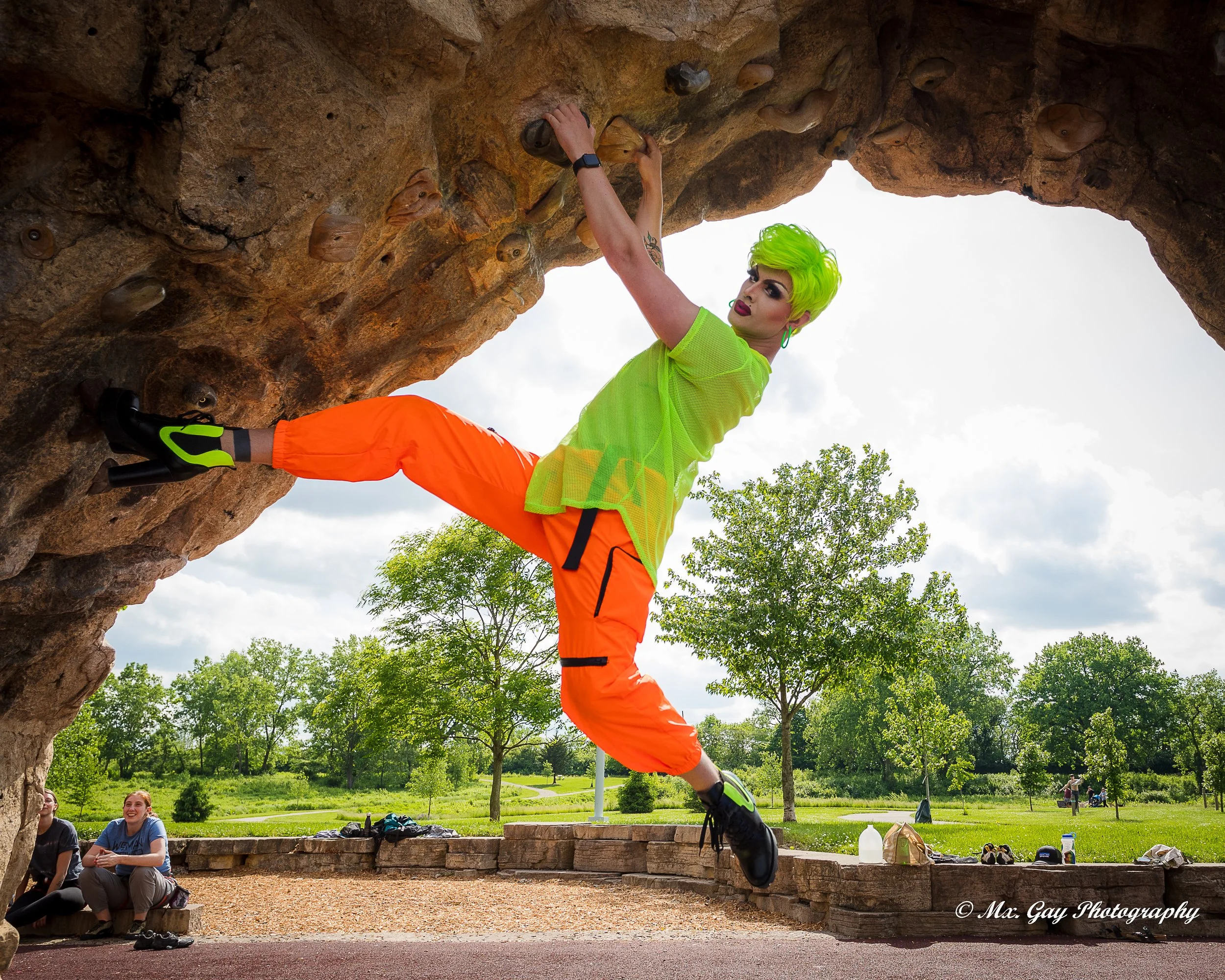 A woman with neon green hair and colorful makeup is climbing a rock formation outdoors, wearing a bright yellow-green shirt and orange pants, with a park and trees in the background.