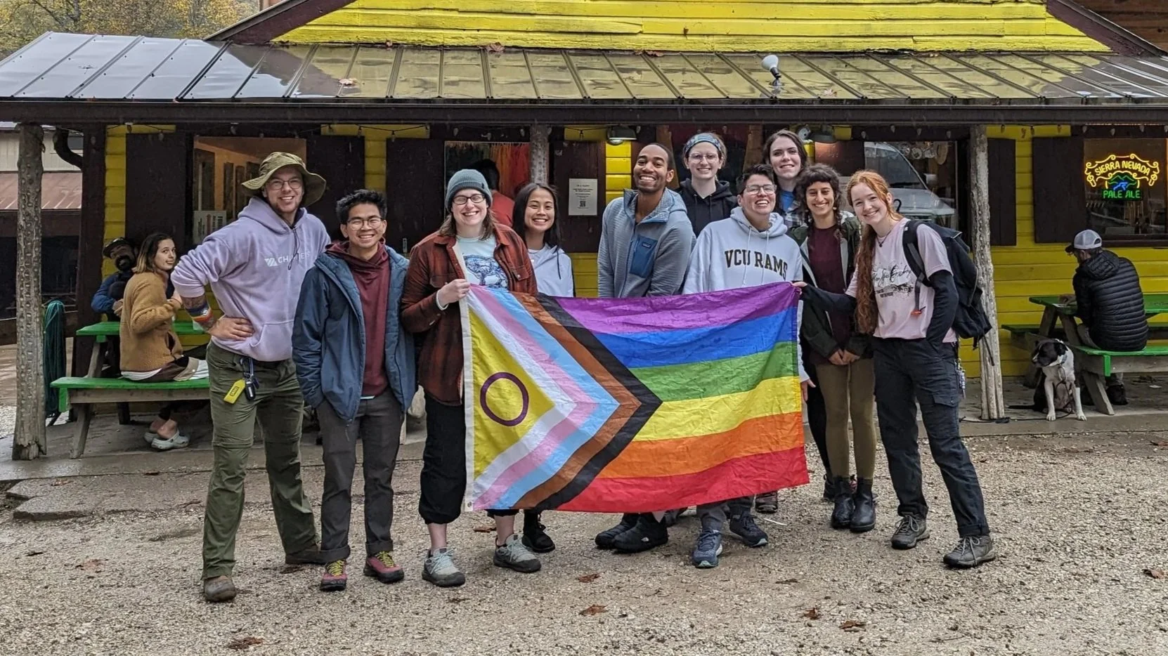 Group of eleven diverse young adults standing outdoors on a gravel surface, holding a rainbow pride flag, in front of a yellow building with a neon sign that reads 'Sierra Nevada Pale Ale' and a green picnic table. Some people are sitting at the table, and one person is holding a dog.