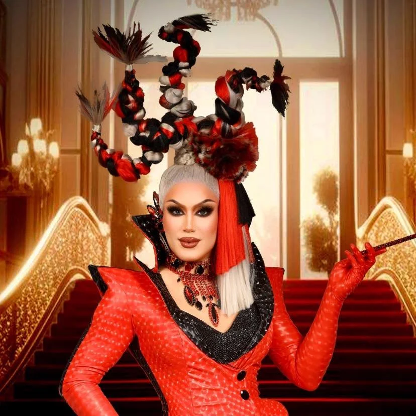 A woman dressed in a red and black costume with elaborate headpiece, standing on a staircase in a luxurious setting.