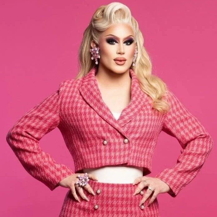 Woman with long blonde hair, wearing pink houndstooth suit with cropped jacket and high-waisted skirt, pink earrings, and large ring, standing against a pink background.