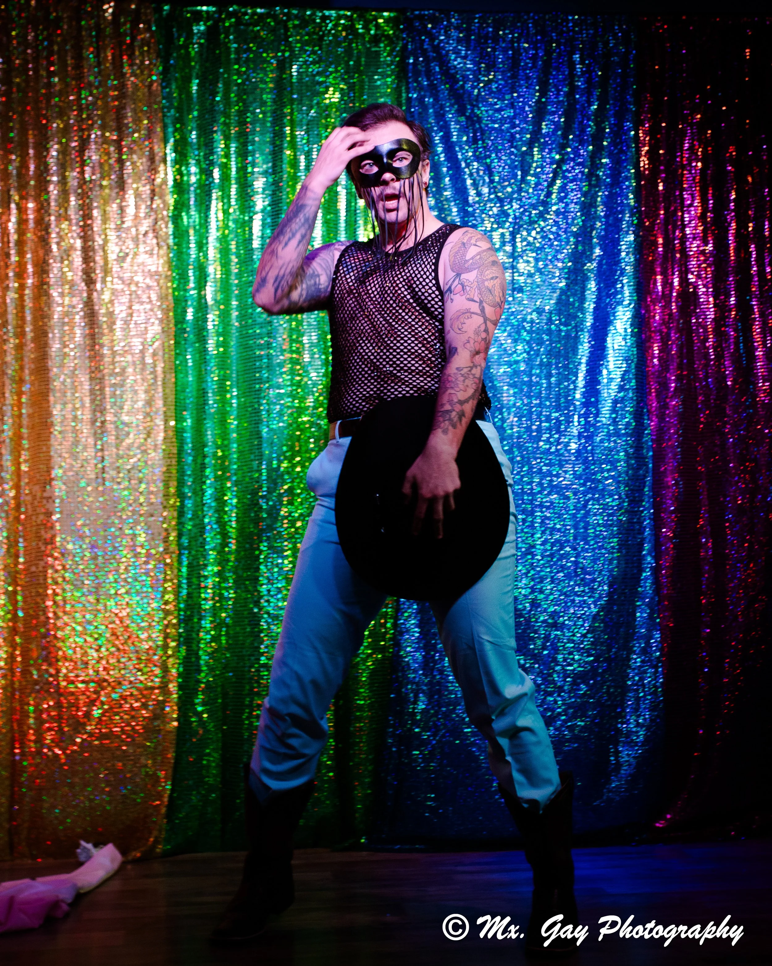 Person wearing a black mask, fishnet top, and light blue pants, standing in front of a colorful glittery backdrop, holding a black hat.