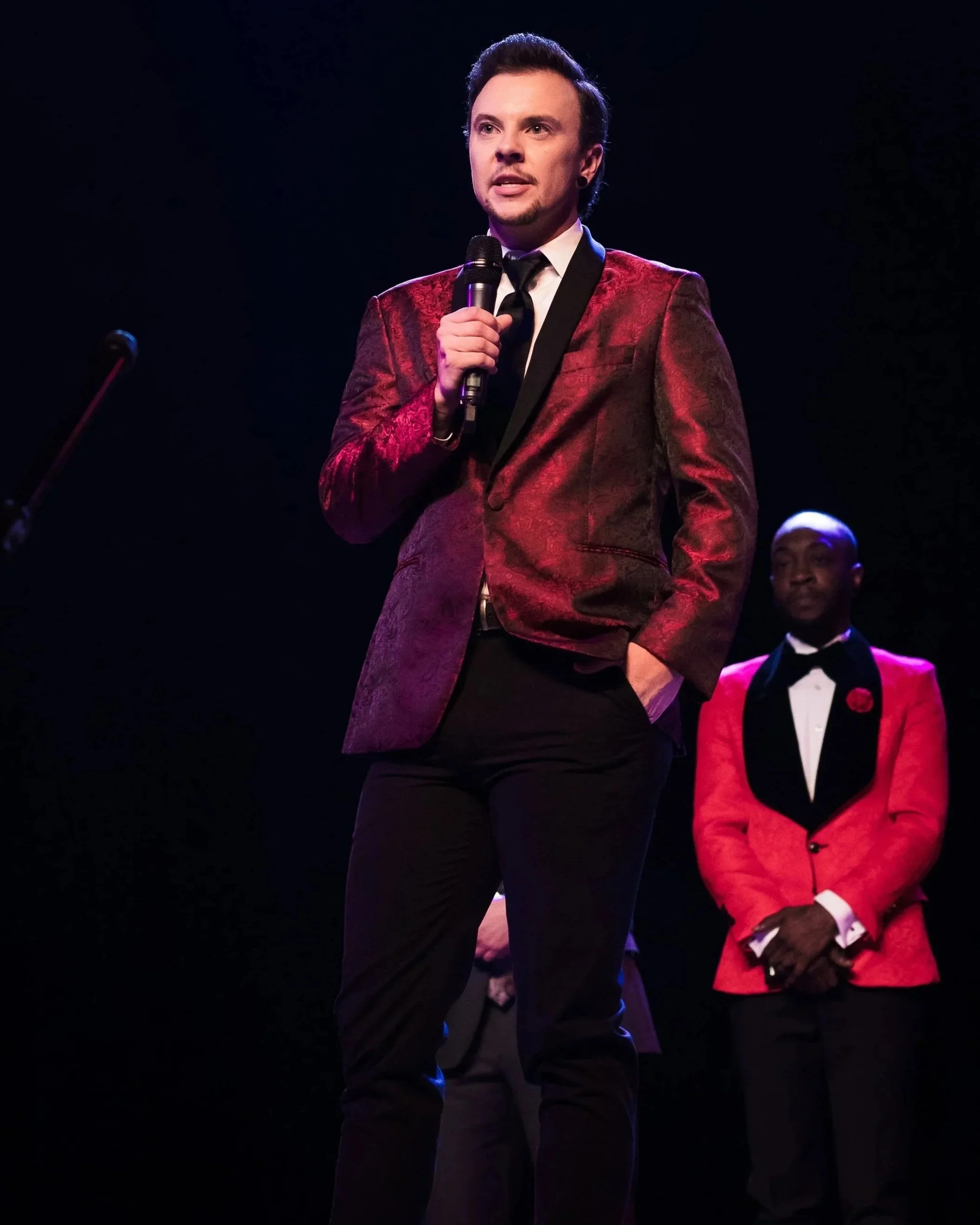 A man in a red patterned blazer, holding a microphone, standing on stage with a dark background. Another man in a red tuxedo with a black lapel and bow tie is visible in the background.