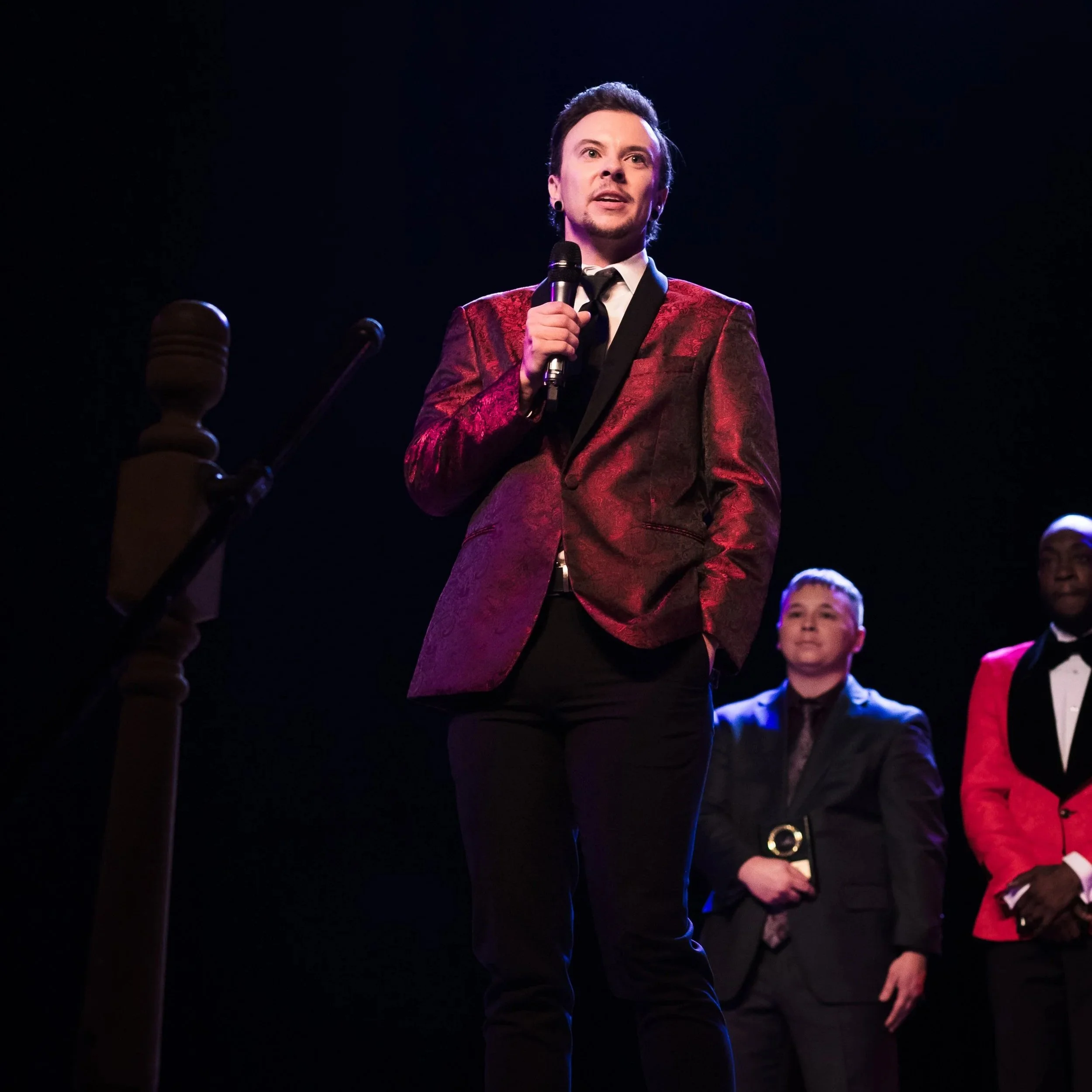 A man in a burgundy suit jacket and black pants speaking into a microphone on stage, with two men in suits behind him.
