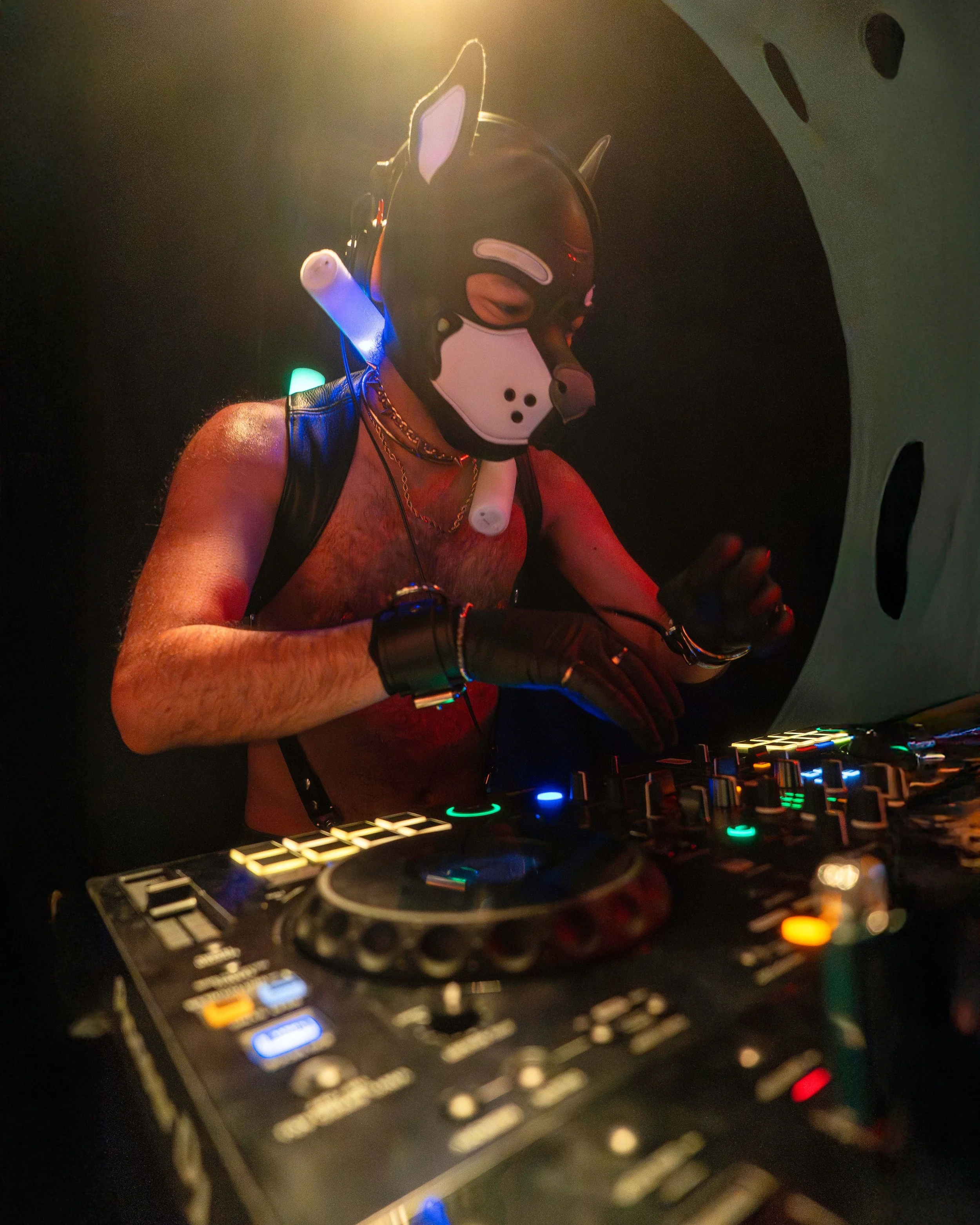 A shirtless DJ wearing a wolf mask with built-in headphones is performing in a dark, atmospheric setting with colorful lights, behind a DJ mixer.
