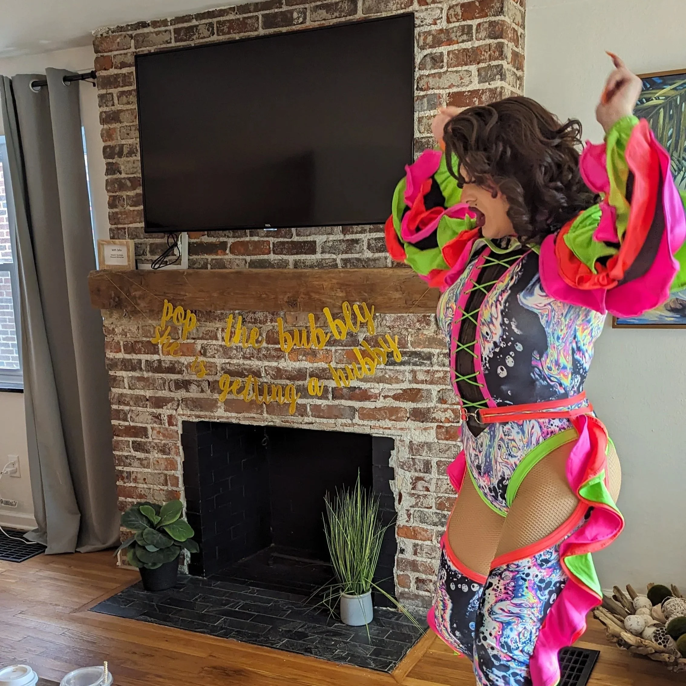 Person dressed in colorful, patterned costume with ruffled pink, green, and black accents, standing inside a living room near a brick fireplace, celebrating and dancing. There are decorative plants and a television above the fireplace, with a sign that says 'pop the bubbly, it's getting a bubbly.'