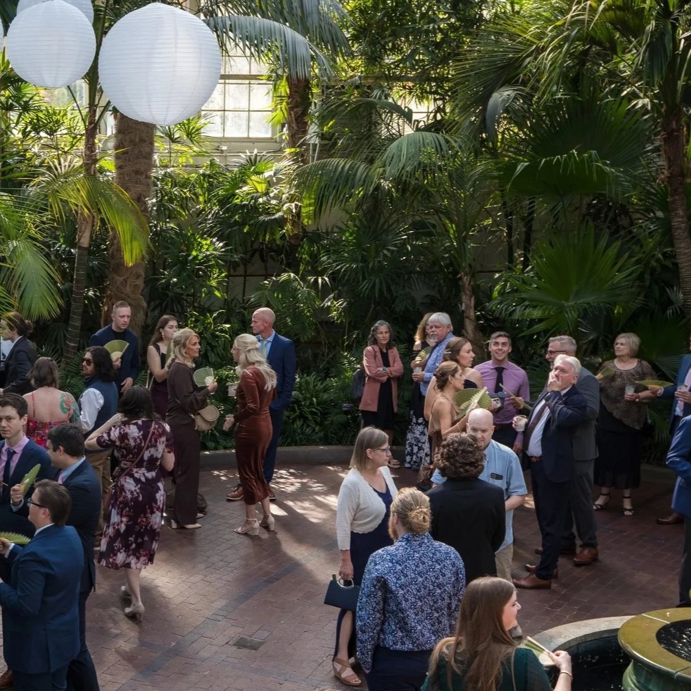 A group of people dressed in semi-formal attire gathered in an indoor tropical garden with lush green plants and large white lanterns hanging from the ceiling.
