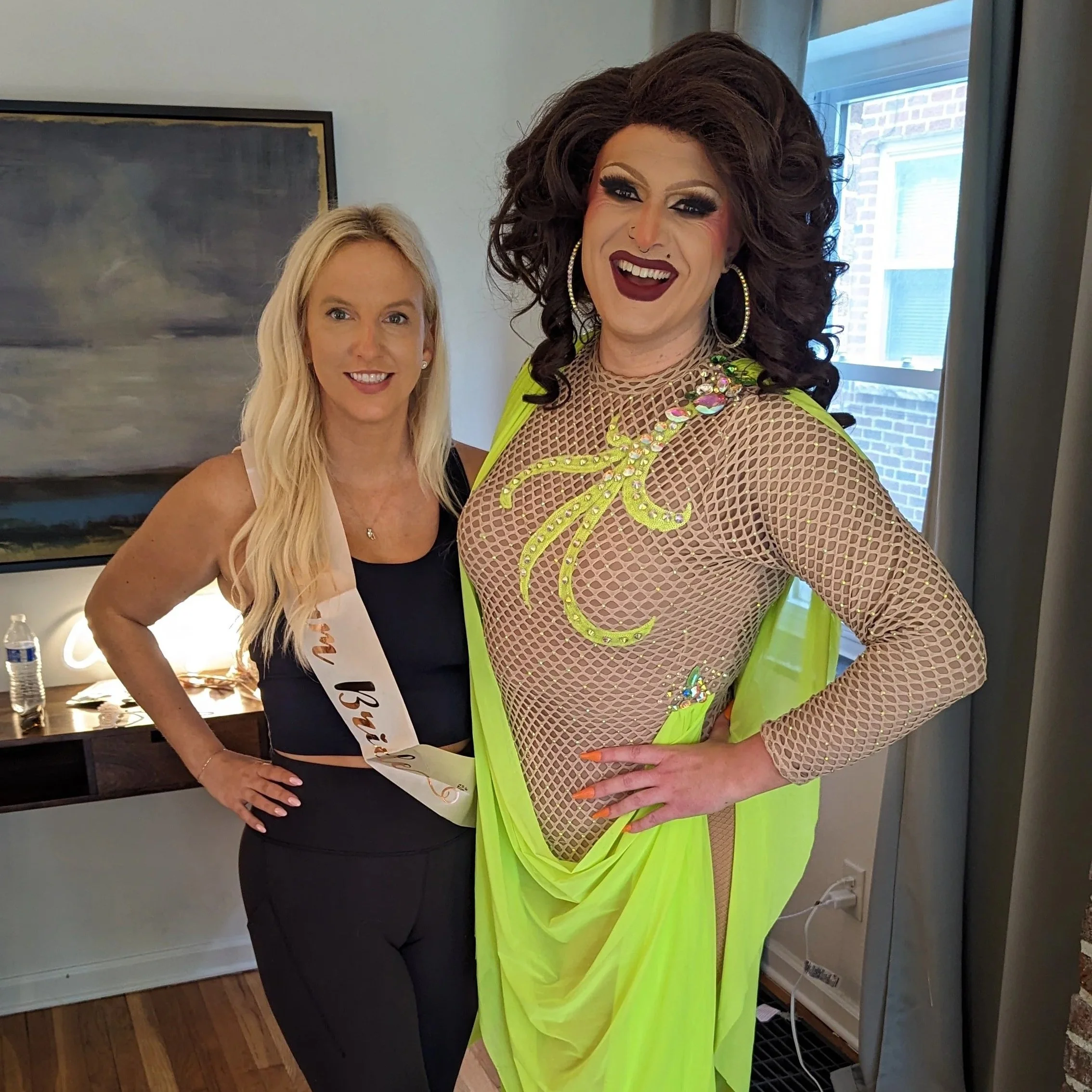 Two women standing indoors, smiling at the camera. One woman is wearing a sash and a black top, and the other is dressed in a yellow and beige outfit with makeup and large earrings.