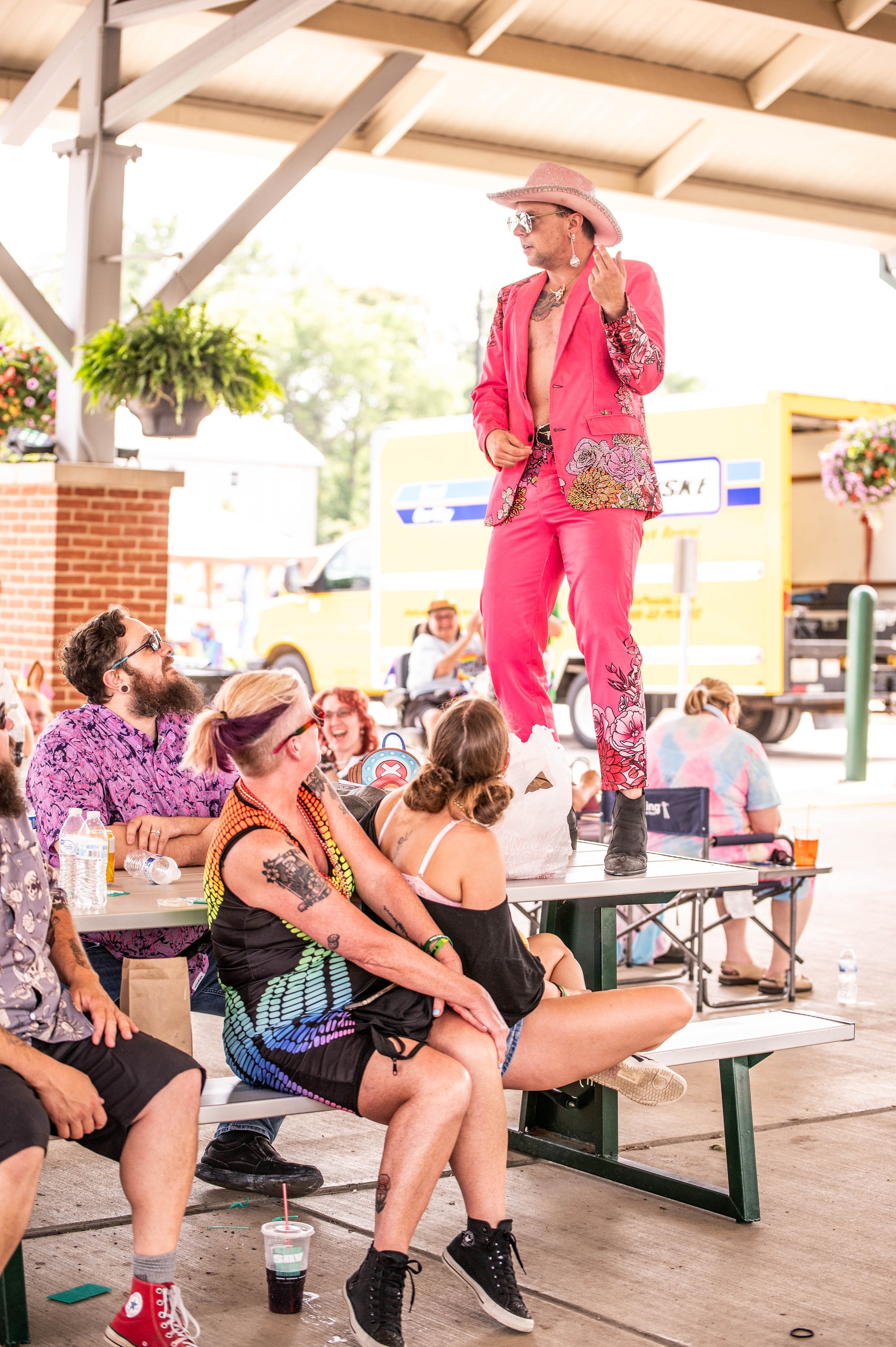 A performer dressed in a bright pink floral suit, pink hat, and dark boots stands on a table under a pavilion, with an audience sitting and laughing, in an outdoor setting during daytime.