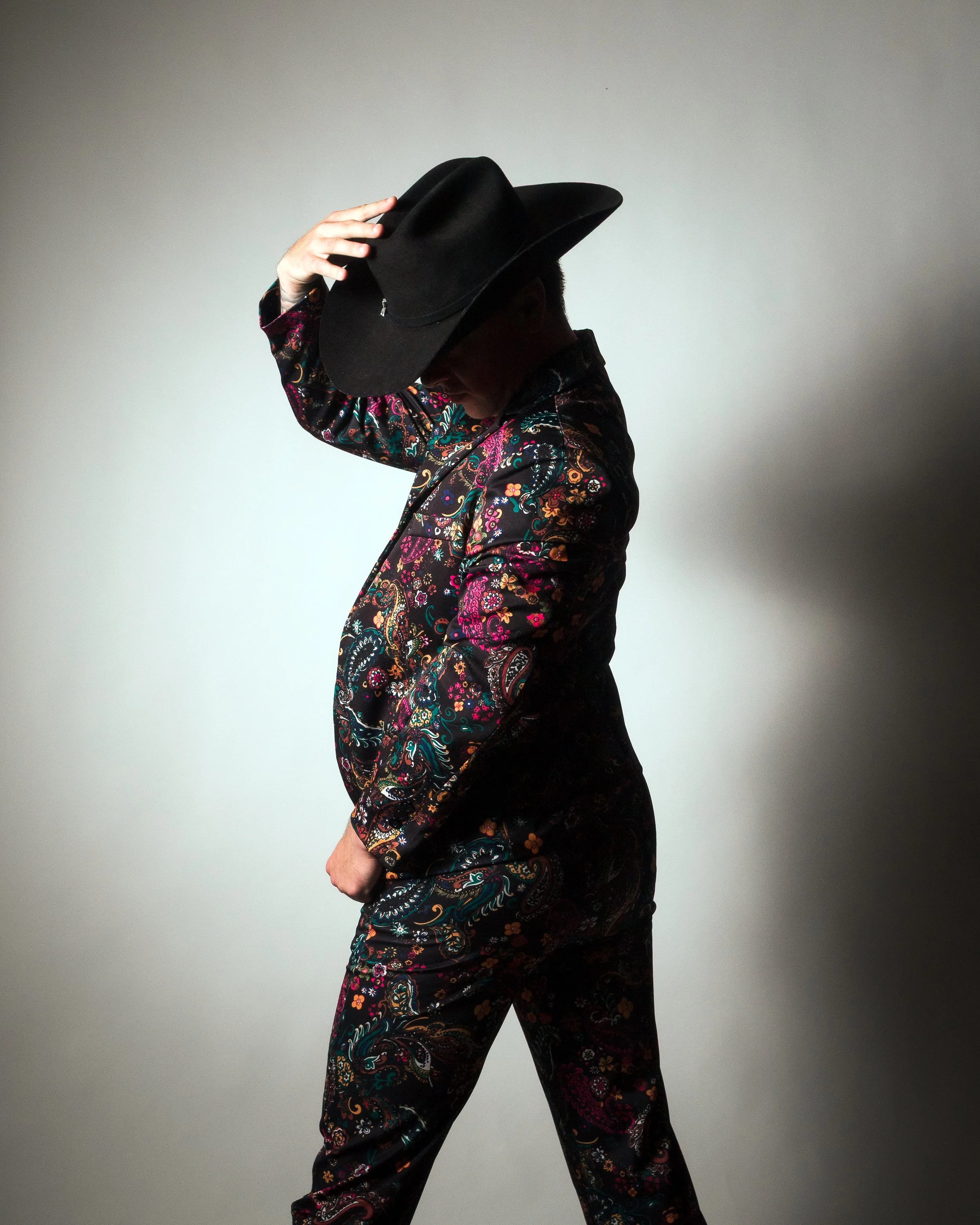 Person wearing a black cowboy hat and a colorful, patterned outfit, posing against a plain background