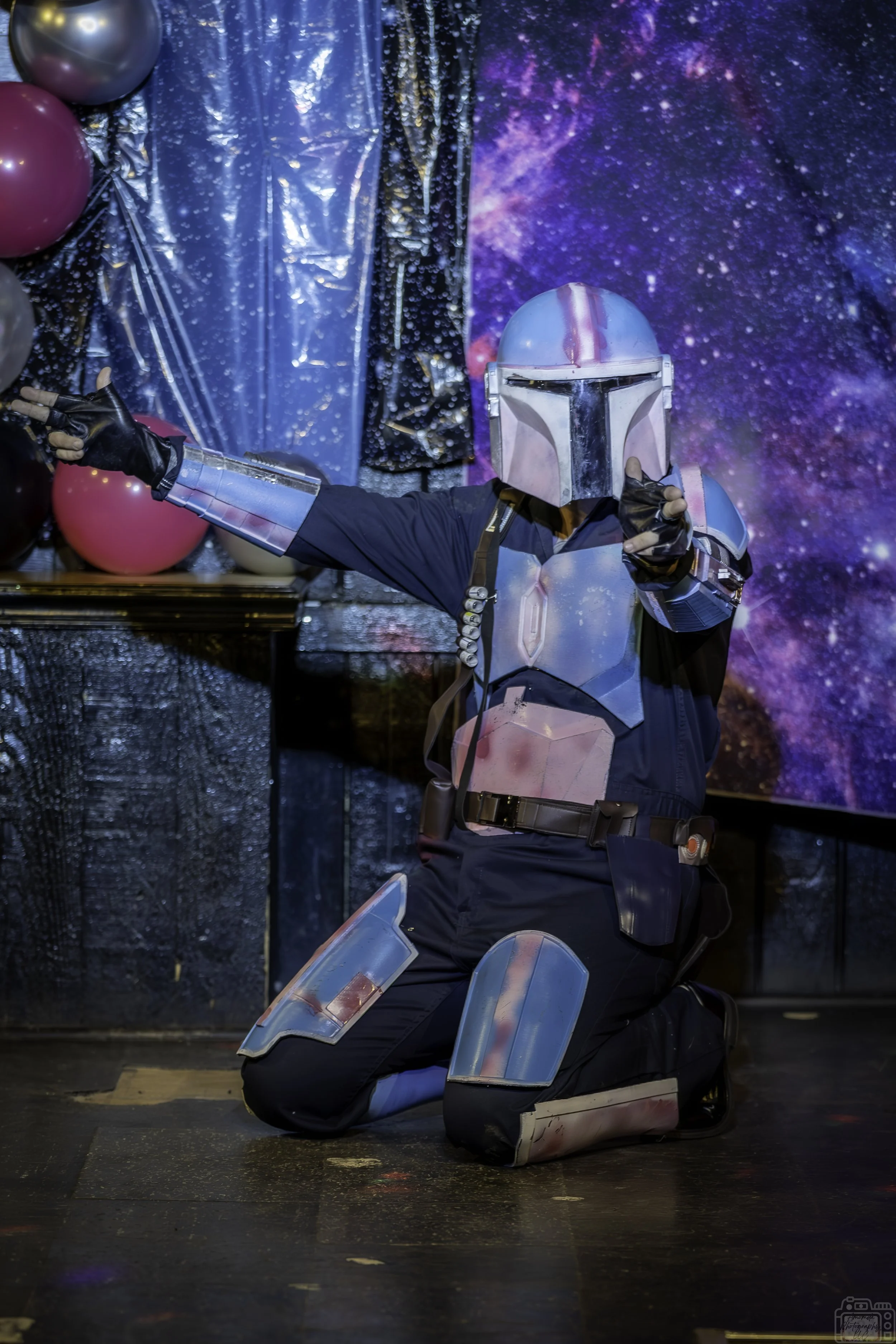 Person dressed as a Mandalorian from Star Wars, kneeling on one knee with a sci-fi background, pointing with one hand, wearing a helmet and armor.