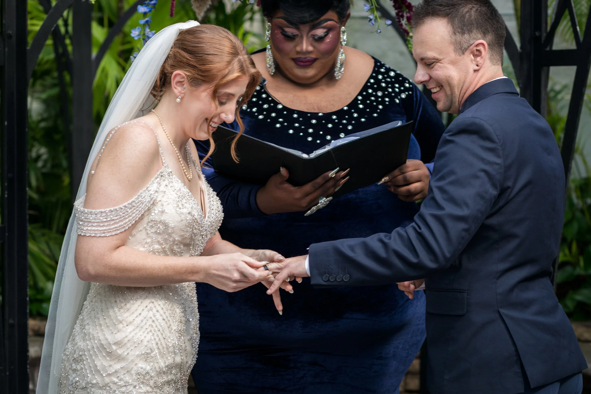 A bride and groom exchange rings during a wedding ceremony, with an officiant reading from a book, in an outdoor setting with green foliage.