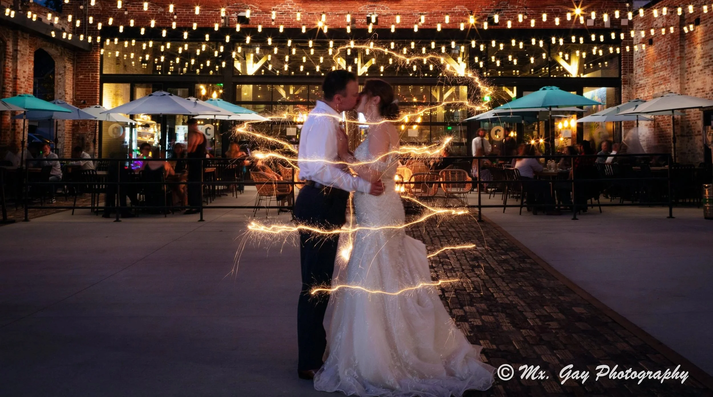 A newlywed couple sharing a kiss and dance at night, surrounded by sparklers, with a venue decorated with string lights and people sitting at tables in the background.