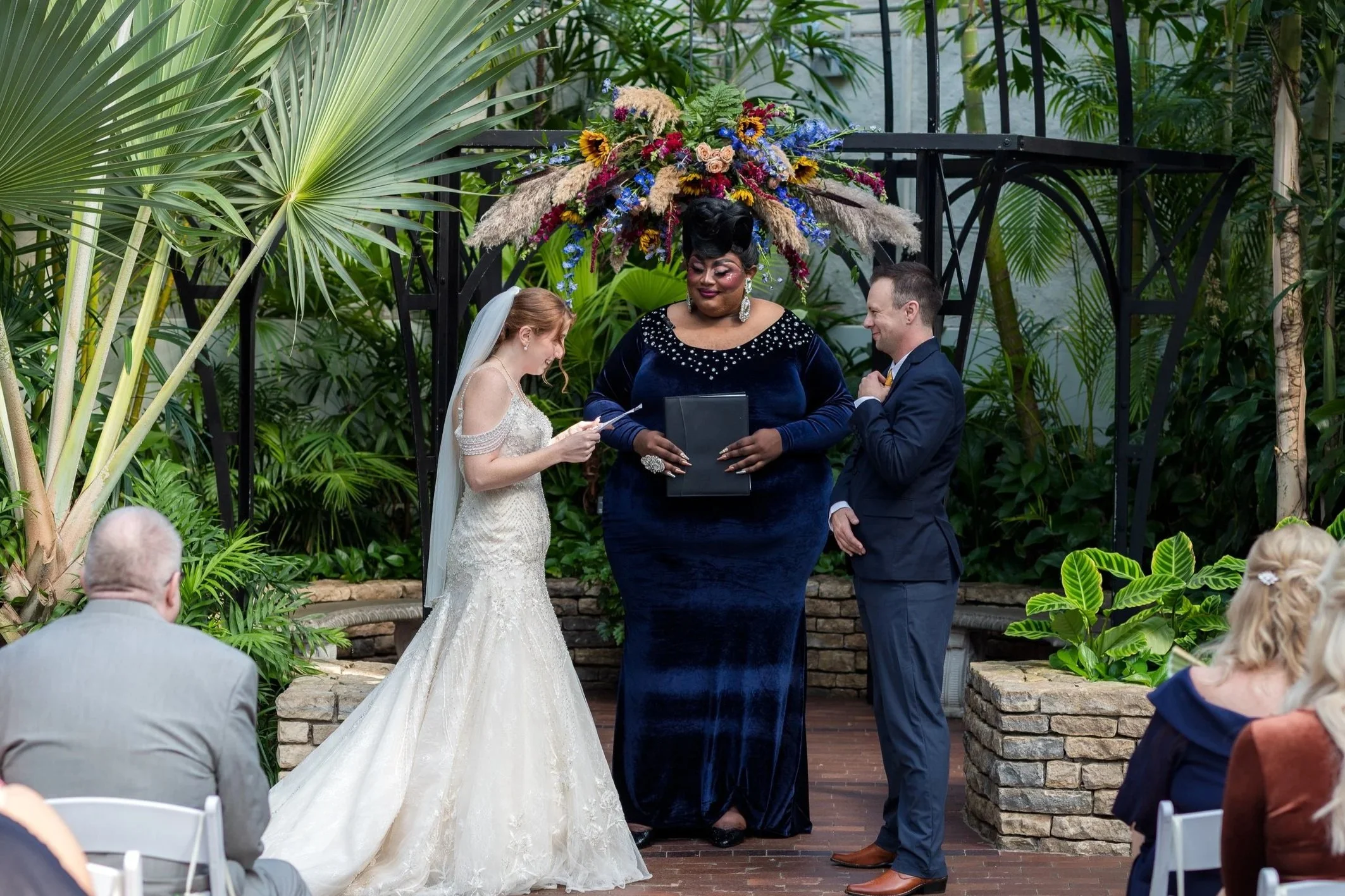 A wedding ceremony taking place in a lush indoor garden setting, with a bride and groom standing in front of a officiant, who is holding a tablet. The bride is in a white lace wedding dress with a veil, and the groom is in a dark suit. The officiant is dressed in a dark blue velvet gown with floral accents on her head and behind her. Guests are seated around, watching the ceremony.