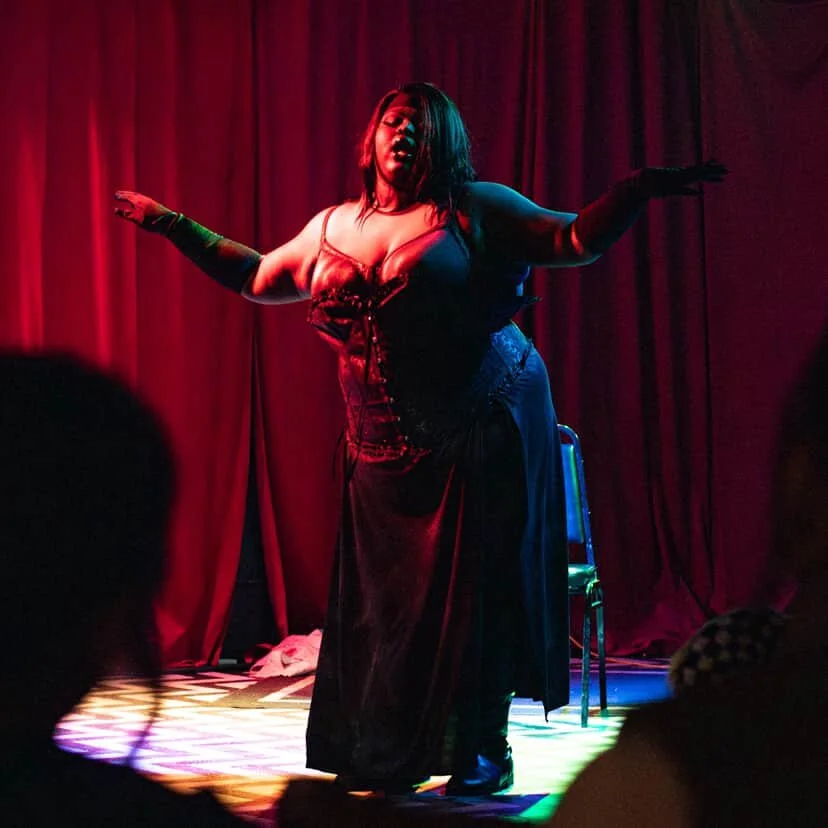A woman performing on stage with red curtains behind her, wearing a black dress and gloves, illuminated by colored stage lights.
