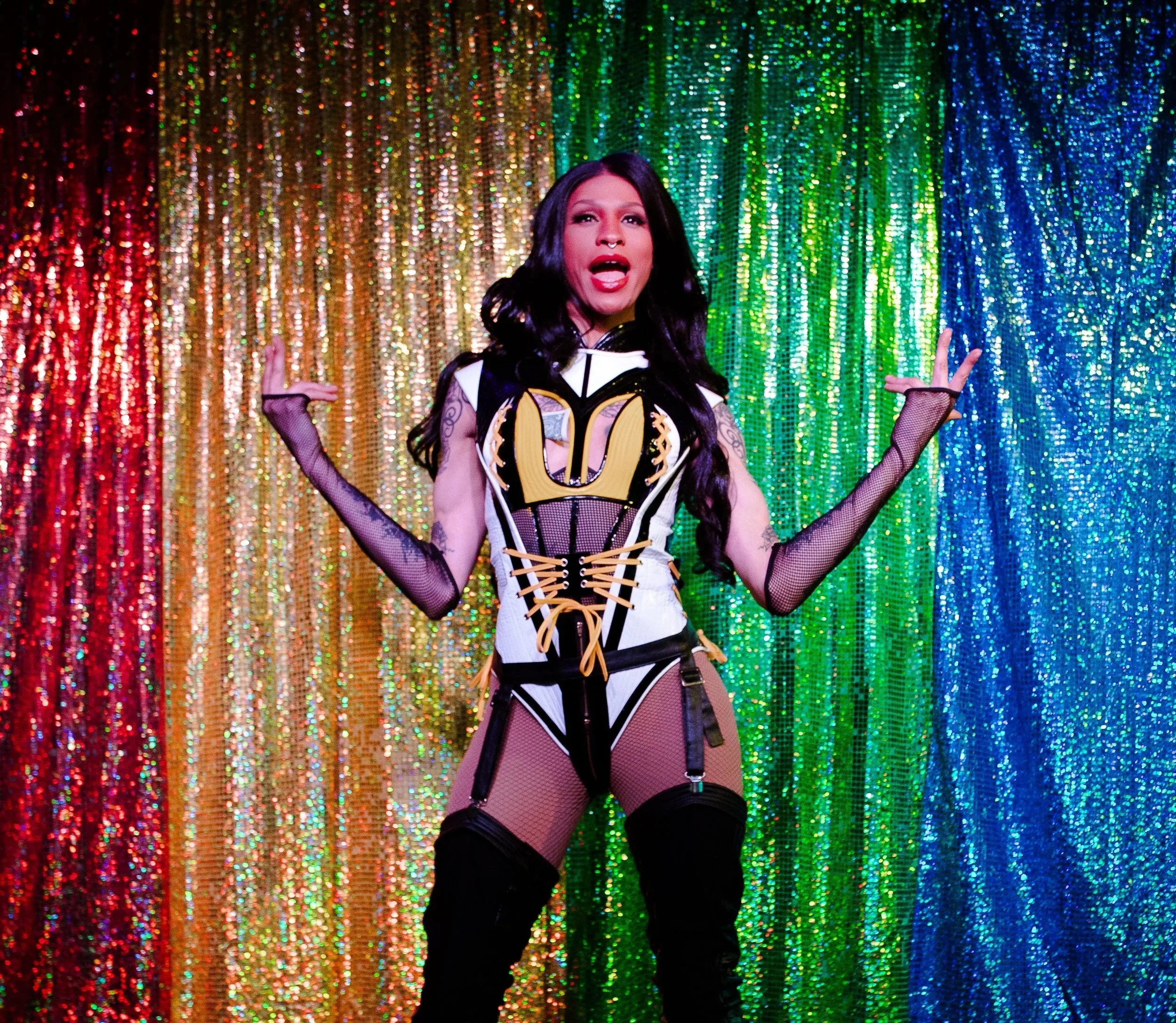 Performer in a colorful, glittery rainbow background, wearing a black and white gothic-inspired outfit with fishnet gloves and thigh-high boots.