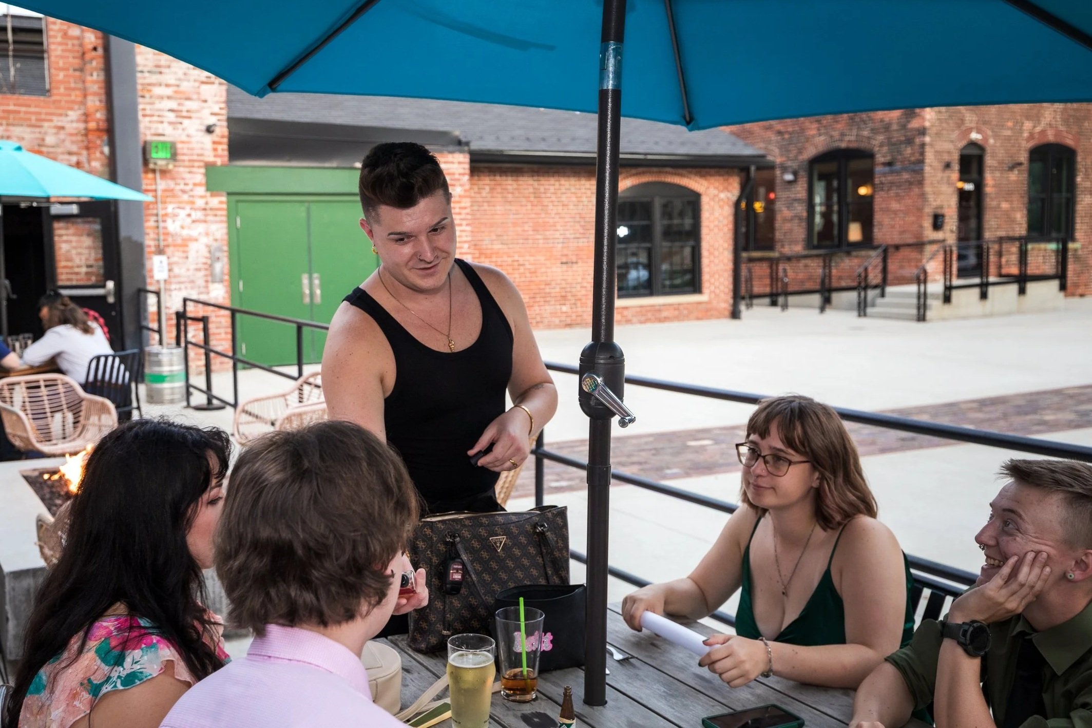 Group of friends sitting at an outdoor restaurant table under a large blue umbrella, engaged in conversation with a waitress nearby.