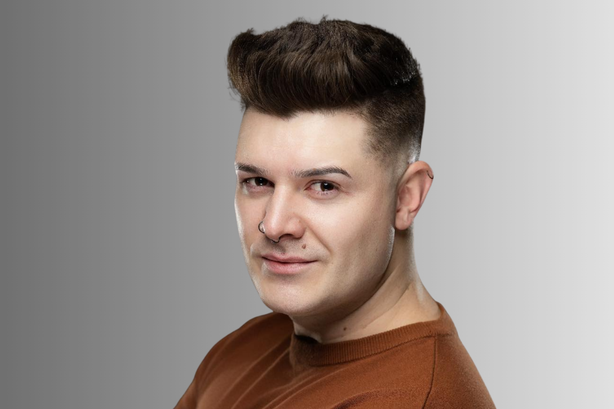 Close-up of a young man with brown hair, light skin, wearing a brown shirt and a septum piercing, smiling softly against a plain gray background.