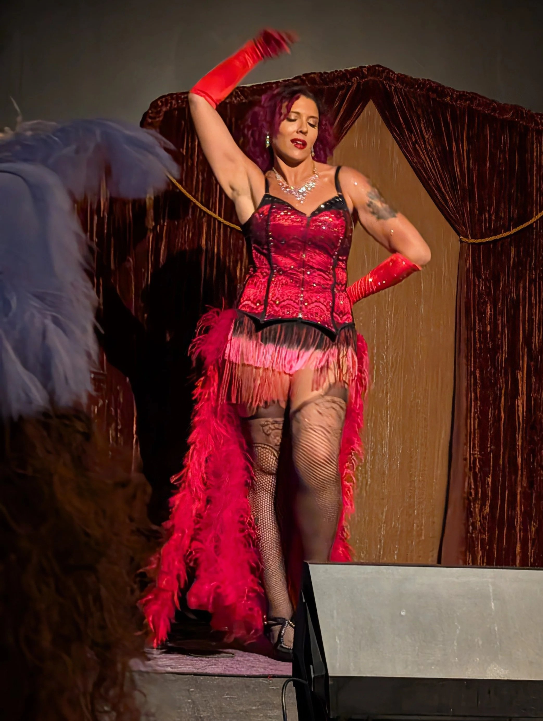 A woman performing on stage in a red and black corset with red feathered skirt, fishnet stockings, red gloves, and high heels, standing in front of velvet curtains.