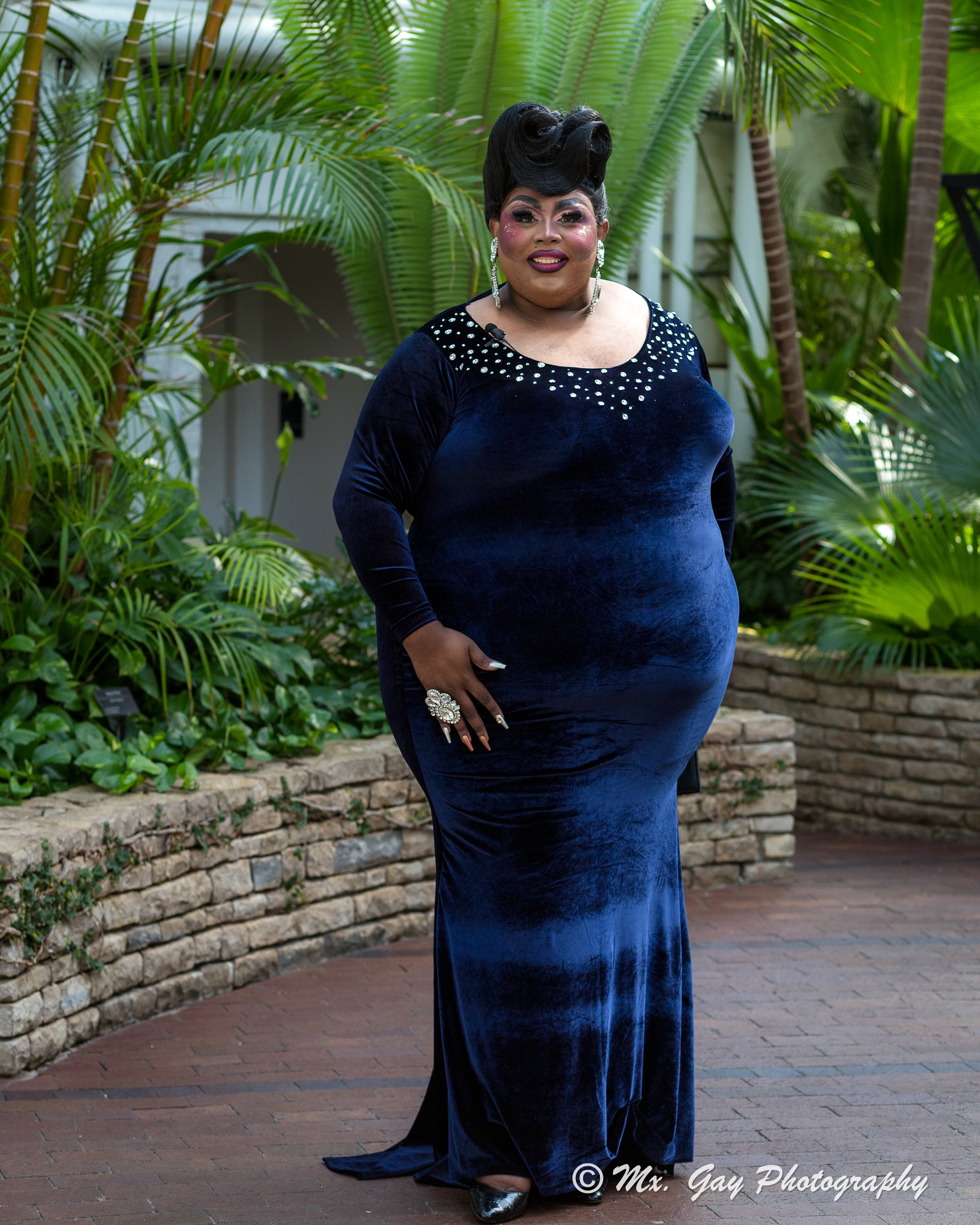 A person dressed in a long, dark blue velvet gown with sparkle embellishments around the neckline, standing outdoors among lush green plants and brick edging.