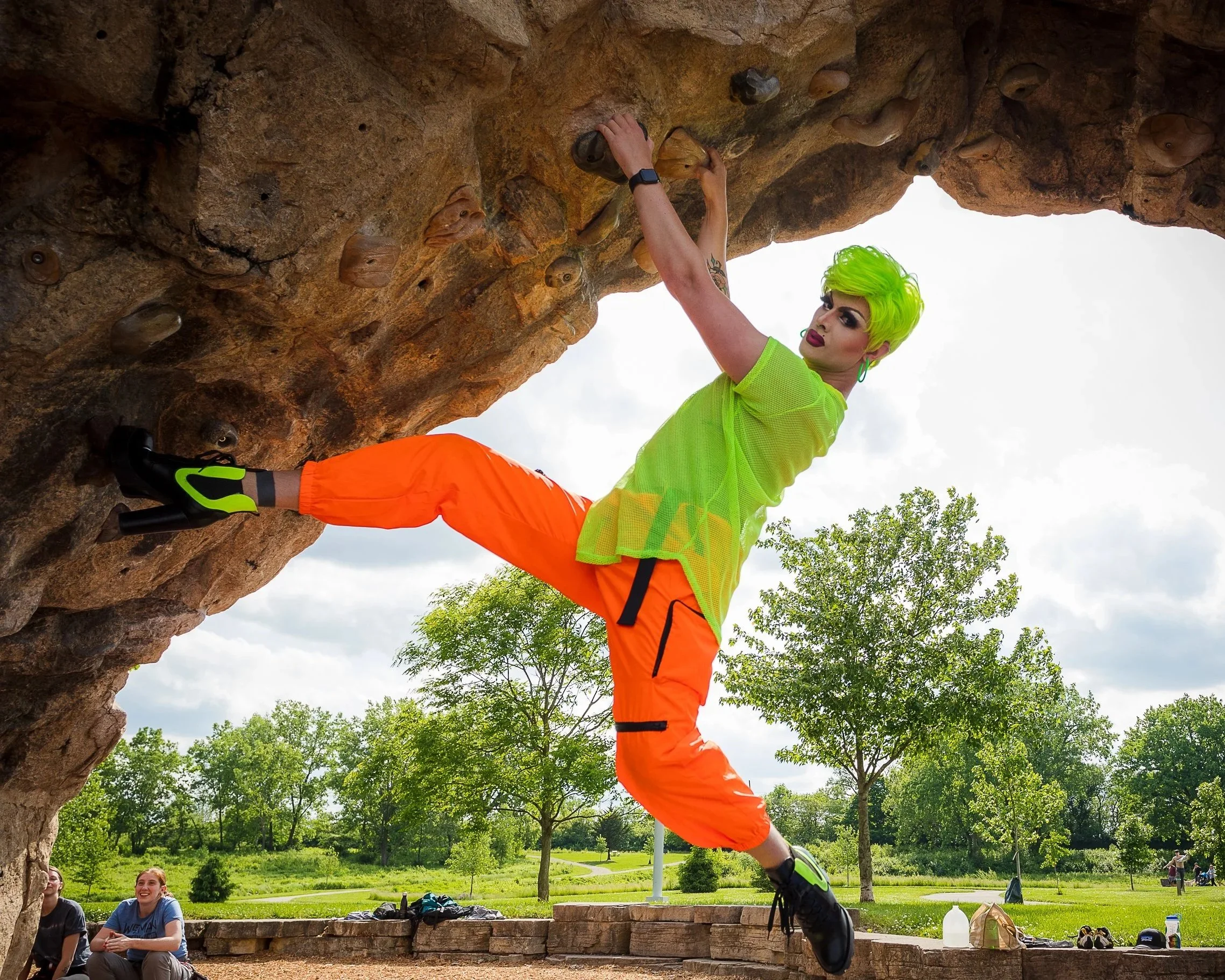 A person with bright lime green hair wearing a neon yellow mesh shirt and orange pants is bouldering on an outdoor rock wall. They are climbing upside down with their feet on the wall and hands gripping holds. Two people are sitting and smiling in the background on the grass, and trees are visible in the distance.