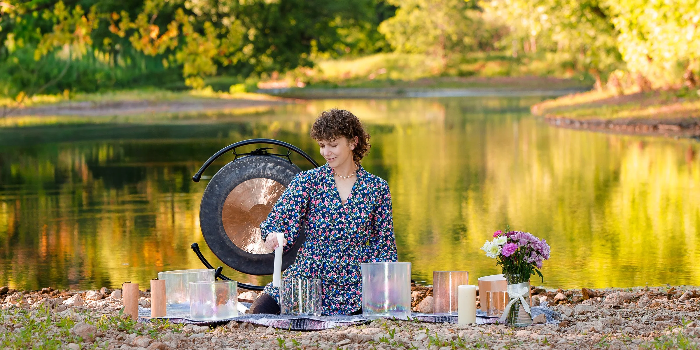 Sound healer plays crystal singing bowls with a gentle, leaf-lined creek behind her.