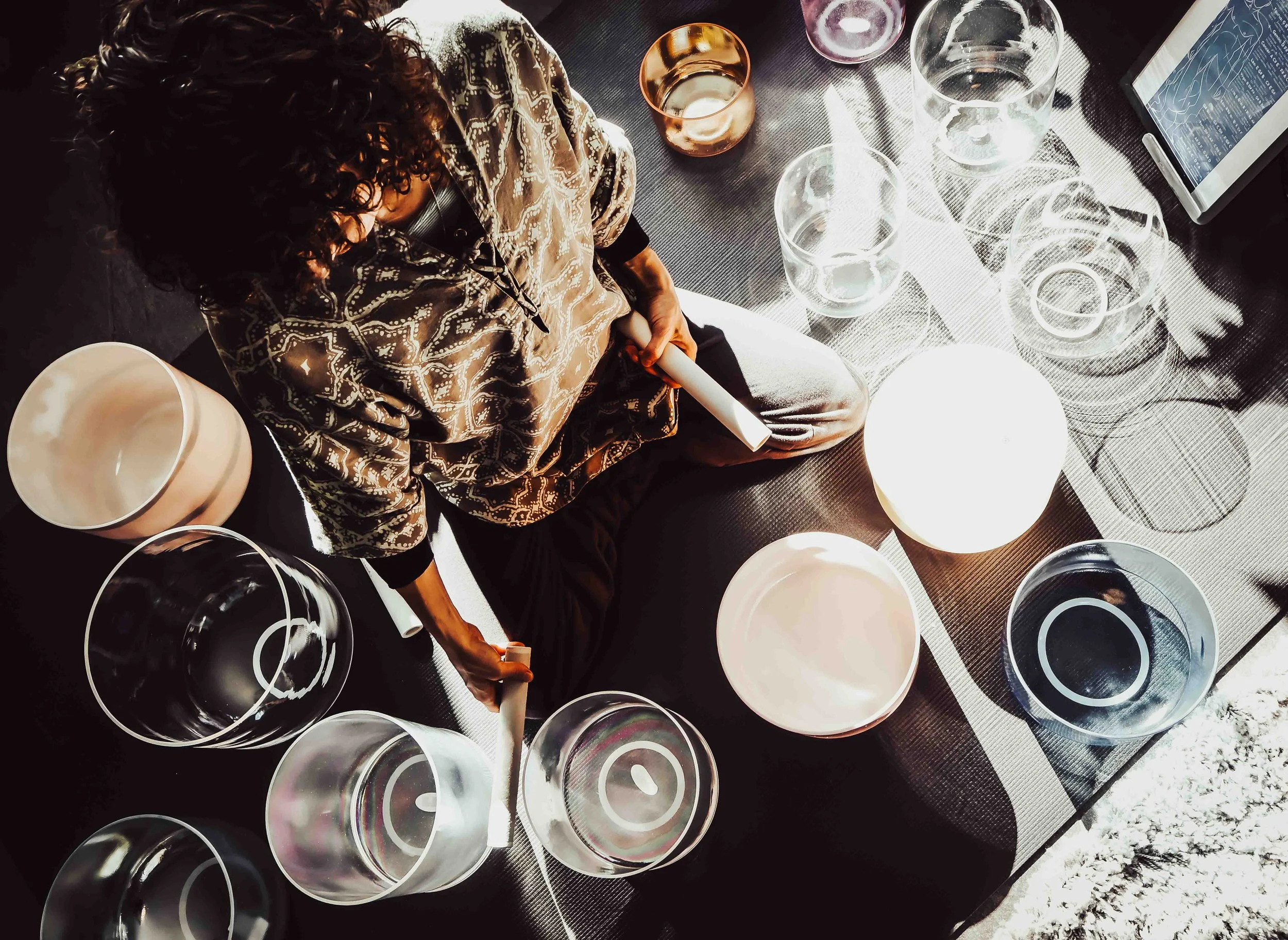 A woman with curly hair wearing a patterned top is sitting at a table with various glassware and dishes. She is holding a rolled-up piece of paper or paper towel. The table has several glasses, pitchers, and bowls, some filled with water. An electronic device with a screen and a stylus is also on the table. Sunlight creates shadows on the tablecloth.