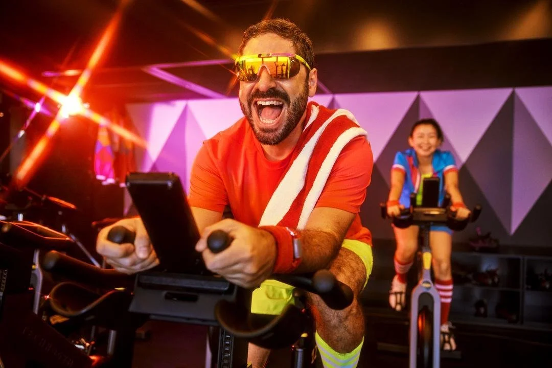 A man riding a stationary exercise bike, smiling and wearing sports glasses and a towel, with a young girl on a similar bike in the background at a colorful fitness studio.