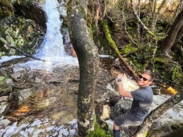 A person wearing sunglasses, a gray t-shirt, and shorts sitting on a tree branch beside a flowing waterfall in a forested area.