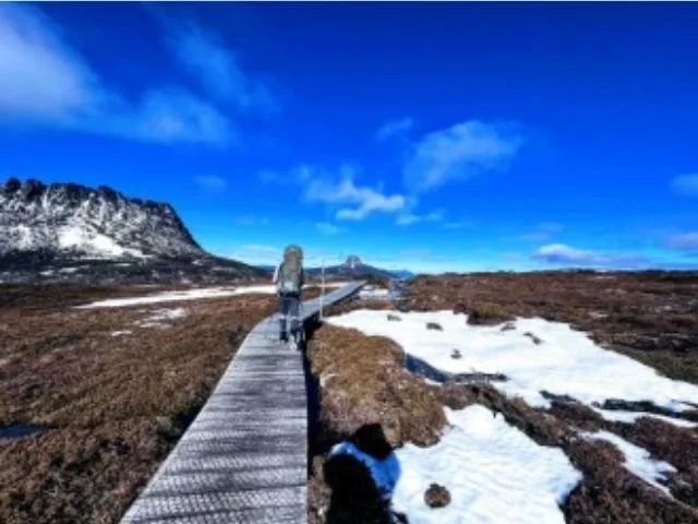 Hiker walking on a wooden trail through a snowy landscape under a blue sky with clouds.