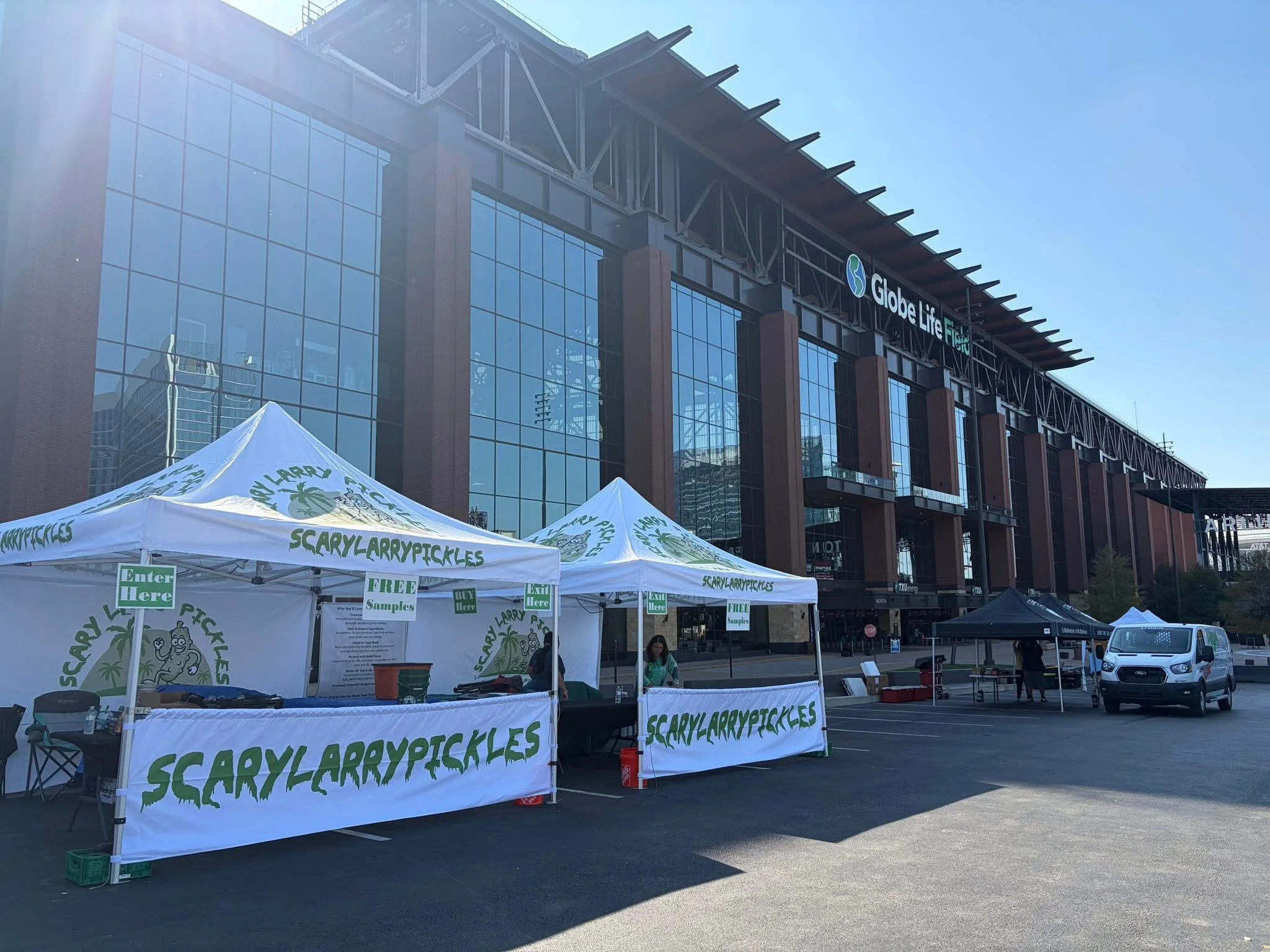 Outdoor market setup with white tents labeled 'ScaryLarryPICKLES' in a parking lot in front of a modern glass and steel building. Nearby are a few people and parked cars.
