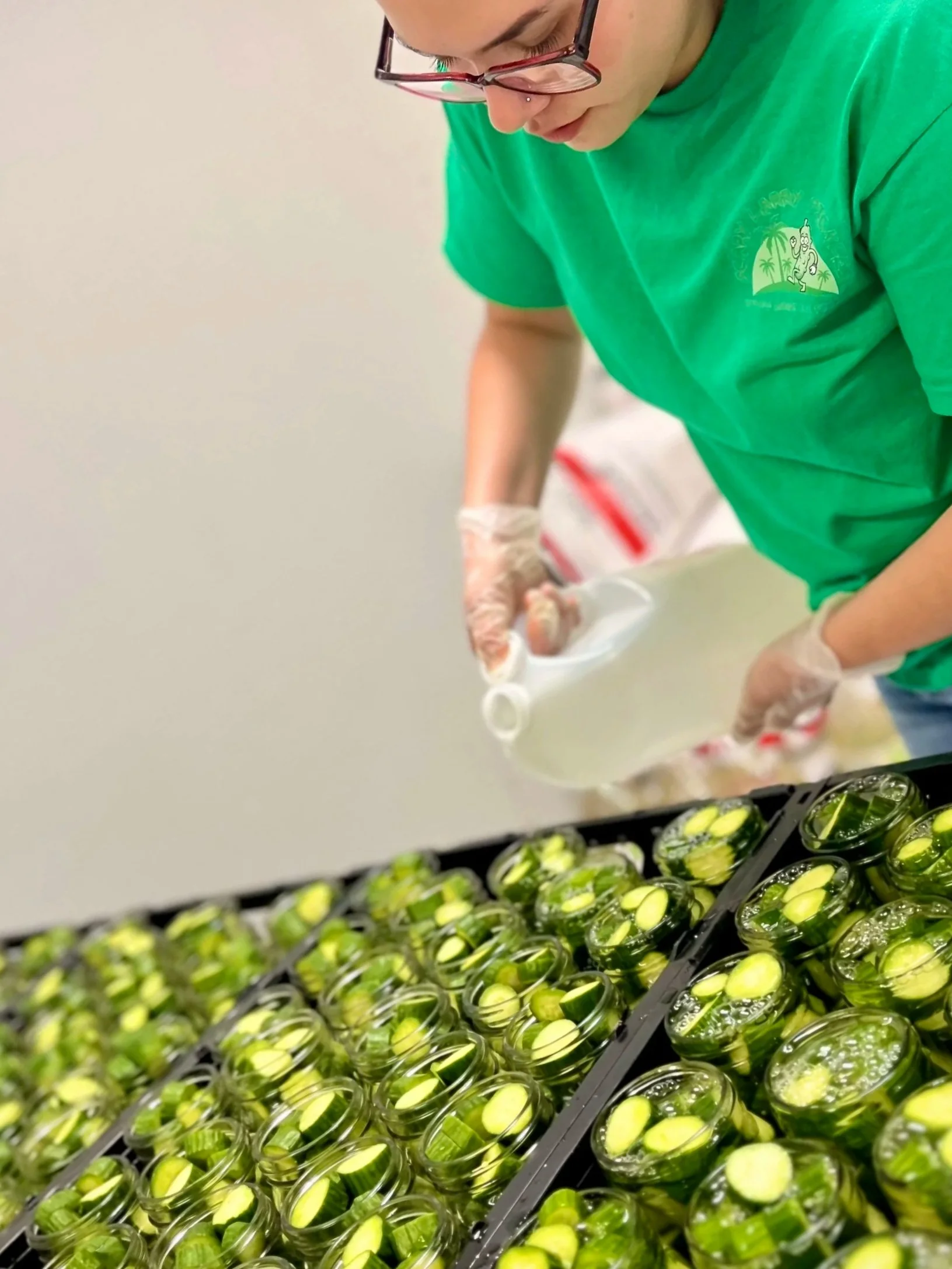 Person in a green shirt and glasses preparing sushi rolls with cucumber and rice in a food prep area.