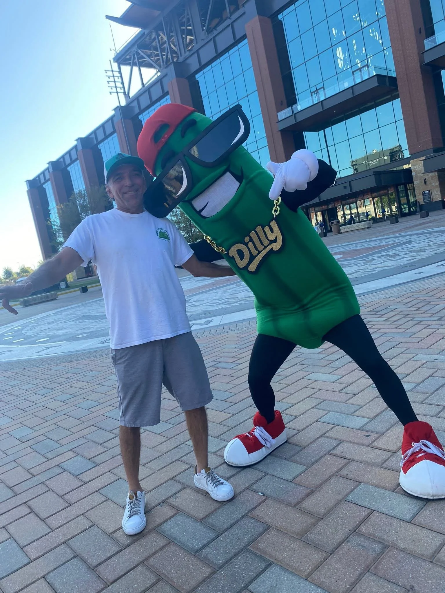 A man standing next to a person dressed in a giant pickle costume with sunglasses, a red hat, and a gold chain, posing outdoors in front of a modern glass building.