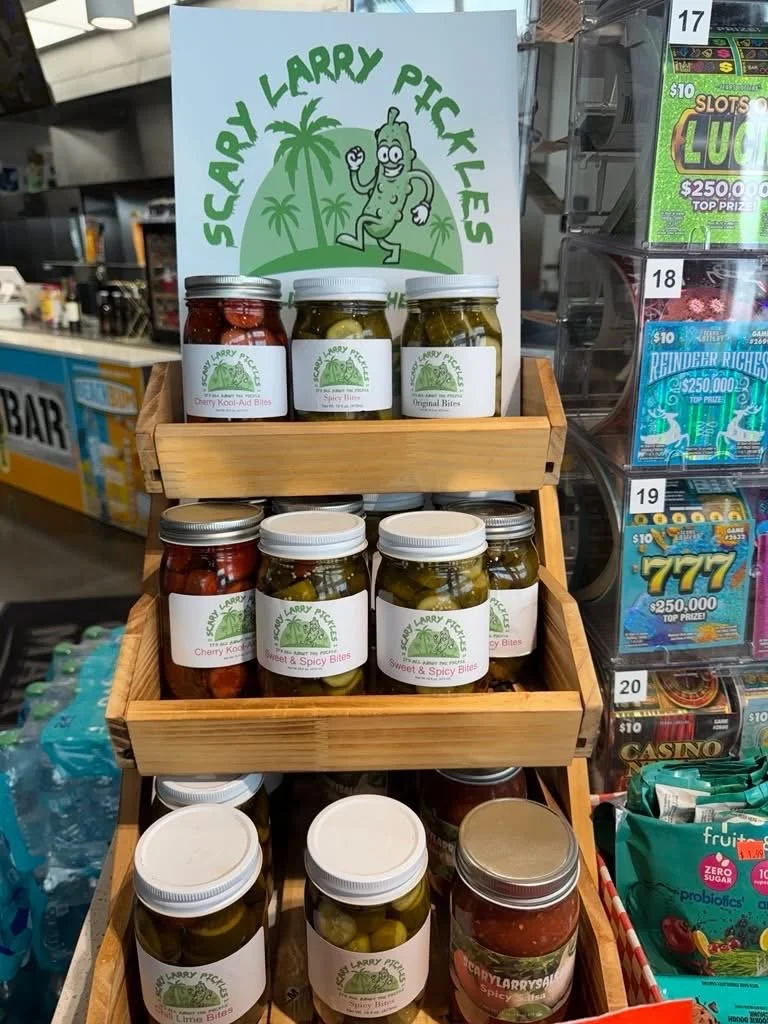 Display stand with jars of pickle slices from Scharly Larry Pickles, including Cherry Kool-Aid Bites, Spicy Bites, Original Bites, and Sweet & Spicy Bites, in a store aisle.