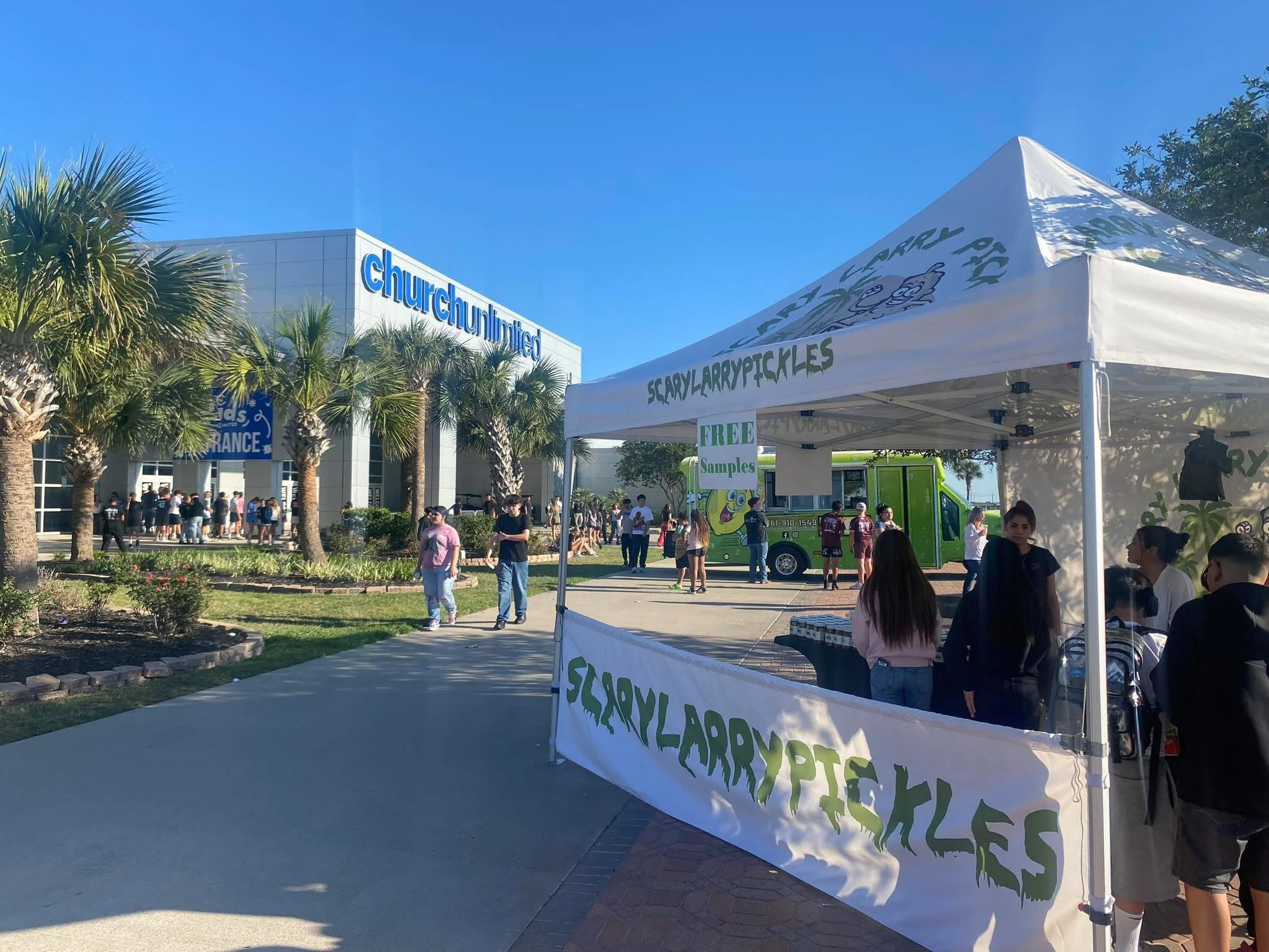 People walking outside near a building with a sign that reads 'churchunited'. There is a white tent with green lettering advertising free samples, and a green food truck in the background. The scene appears to be a community event on a sunny day.