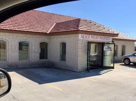 Exterior view of Alice Meat Market store with brick walls, a red tiled roof, and an open door, in a parking lot with a white vehicle parked nearby.