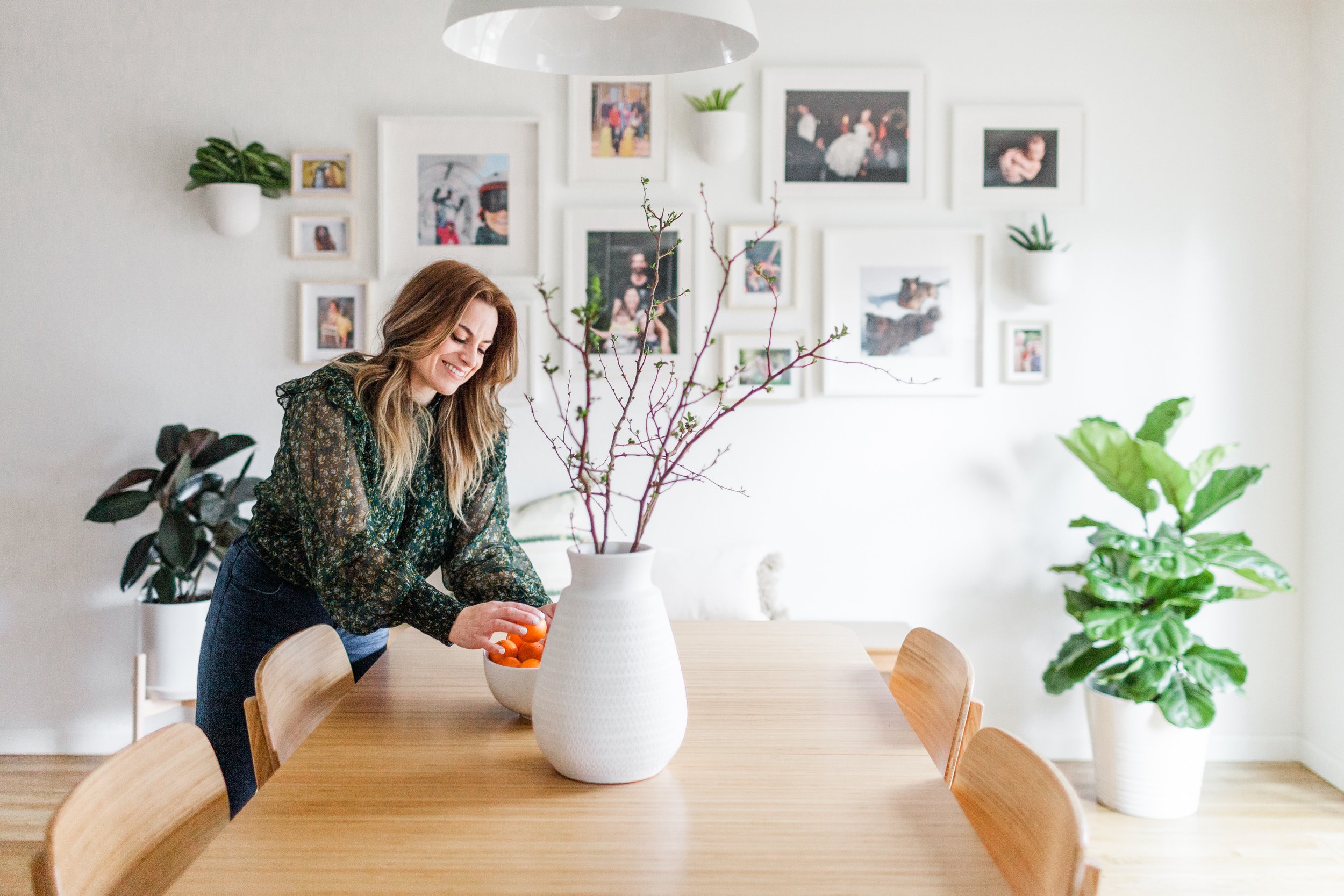 A woman with long wavy blonde hair, wearing a dark floral blouse, stands at a wooden dining table, smiling and reaching into a white bowl filled with oranges. The room is decorated with gallery wall frames containing photos, and there are several potted plants on the wall and table, with soft natural light illuminating the space.