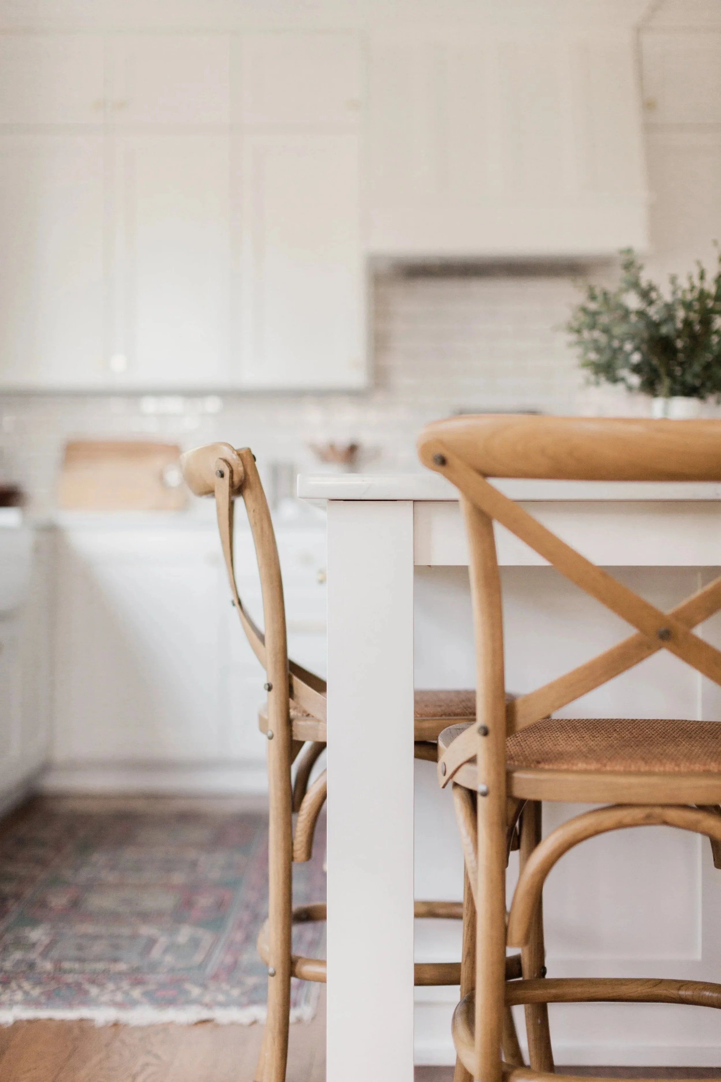 Wooden chairs around a white table in a cozy home dining area with a patterned rug and a potted plant in the background.