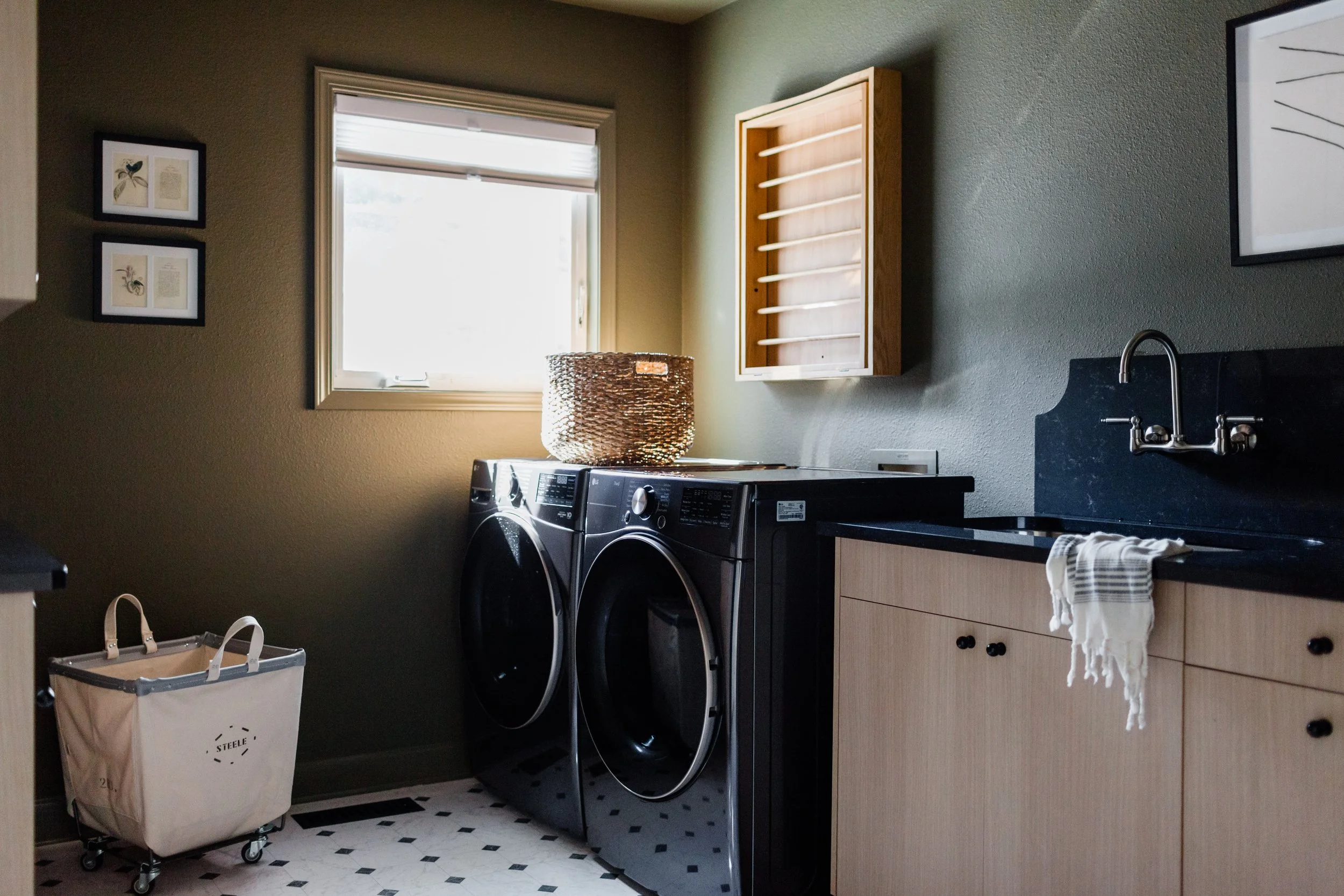 A laundry room with a window, a wicker laundry basket on top of a washing machine, a small cart, and a wood cabinet with open shelves, a sink, and framed artwork on the wall.