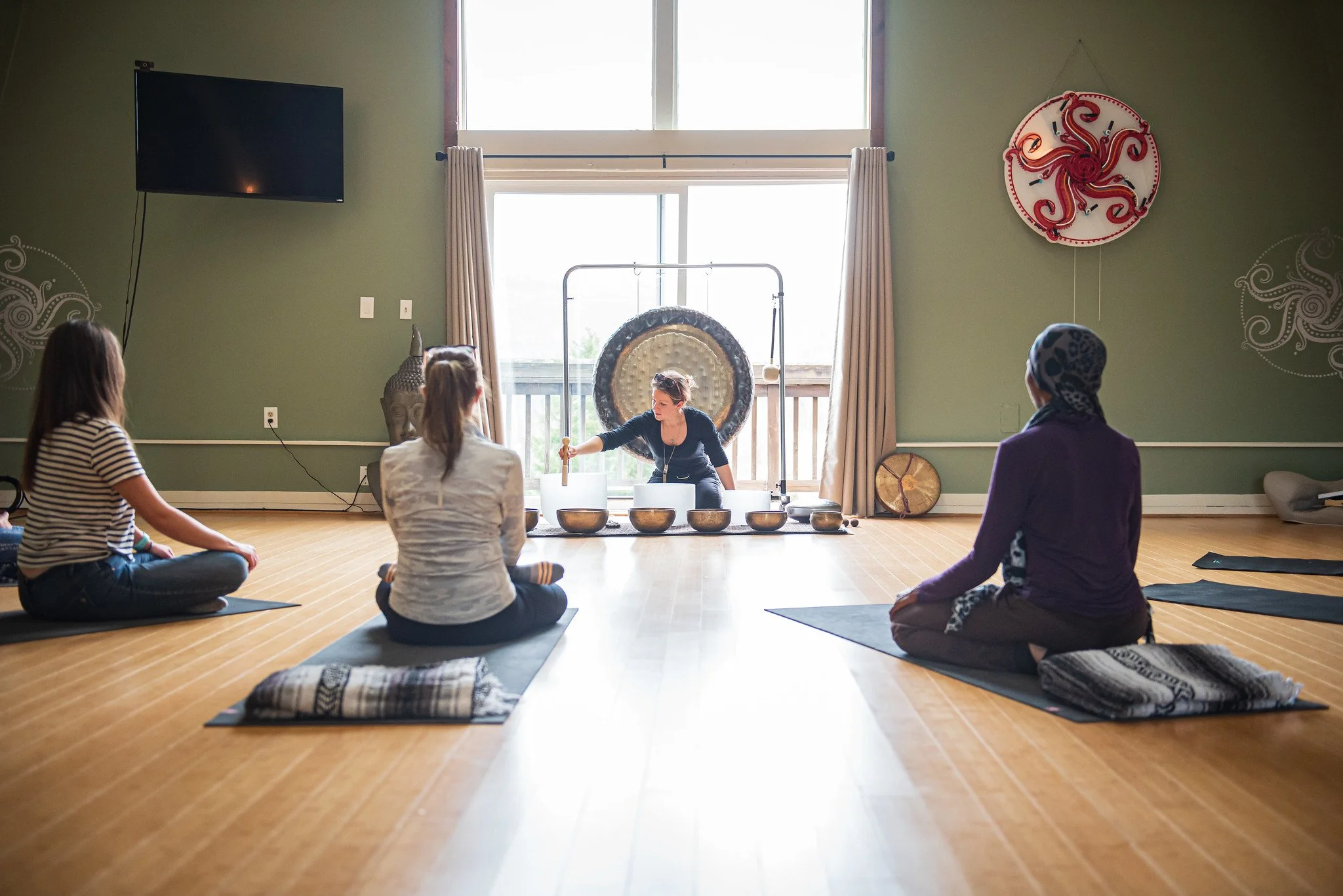A yoga or meditation class with four women sitting cross-legged on mats in a spacious room, facing a instructor who is demonstrating a meditation technique with a gong behind her, natural light coming through large windows.